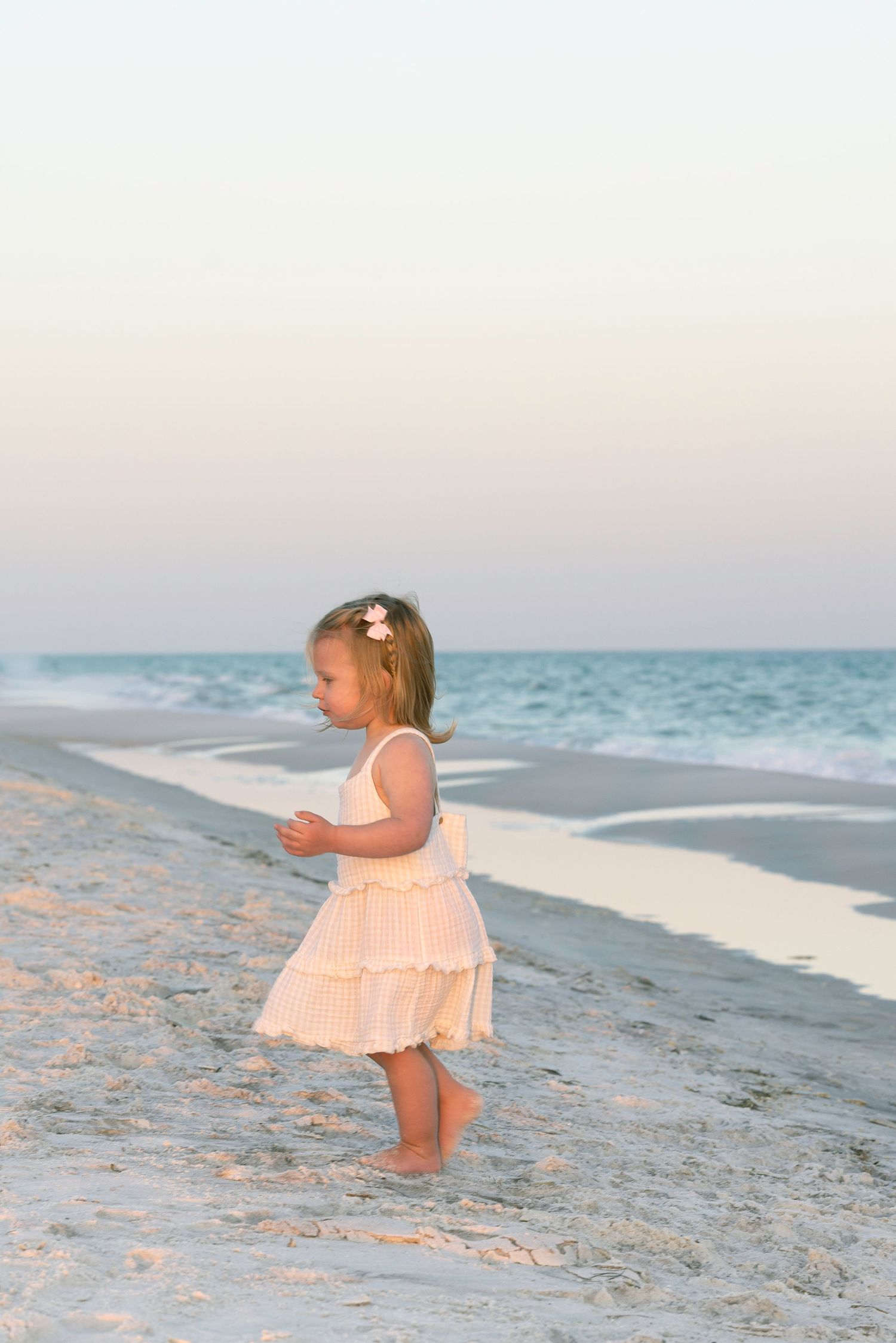 A young toddler girl in a white dress walks barefoot on a sandy beach at sunset.