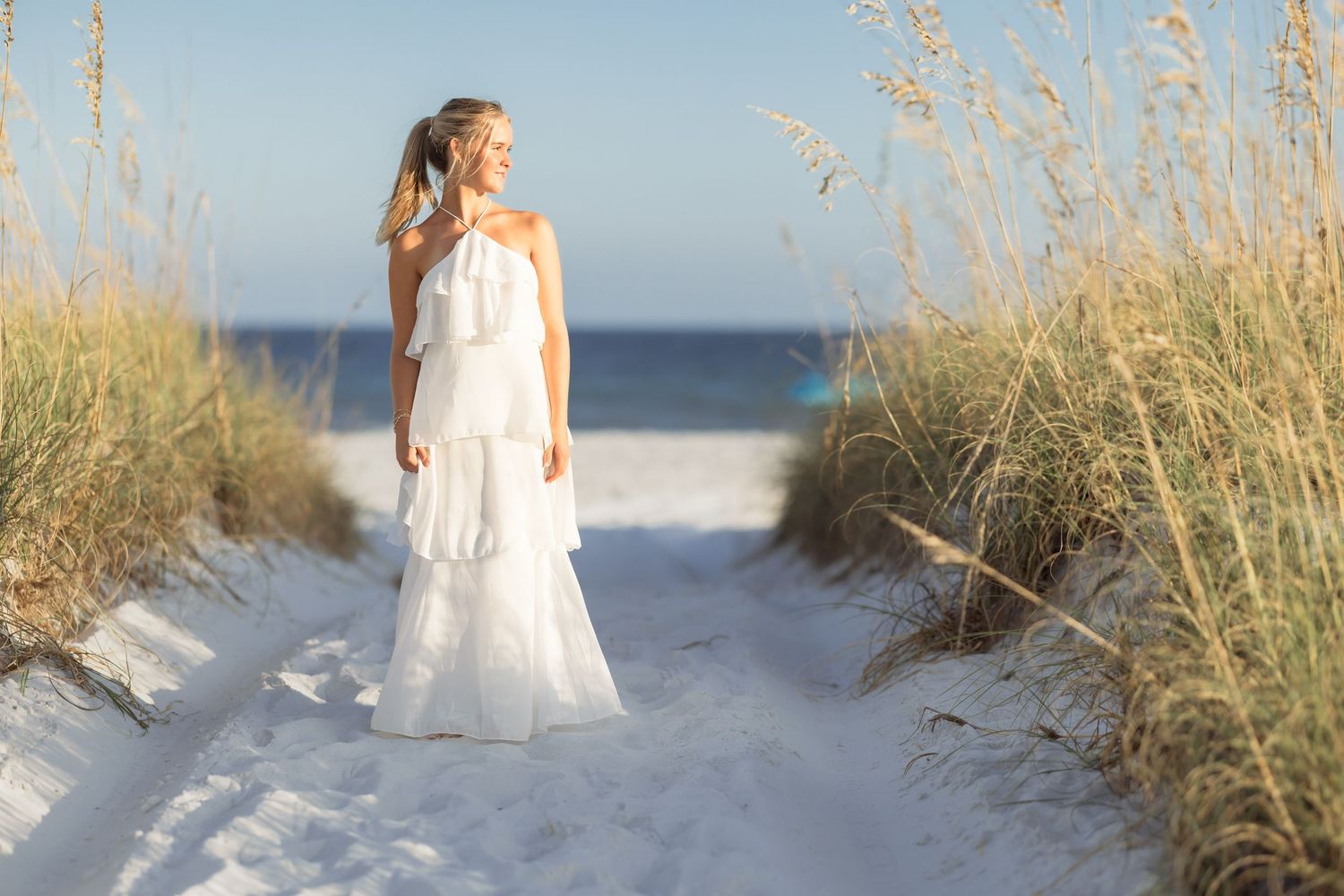 A woman in a white flowing gown stands on sandy beach dunes surrounded by sea oats with ocean in background.