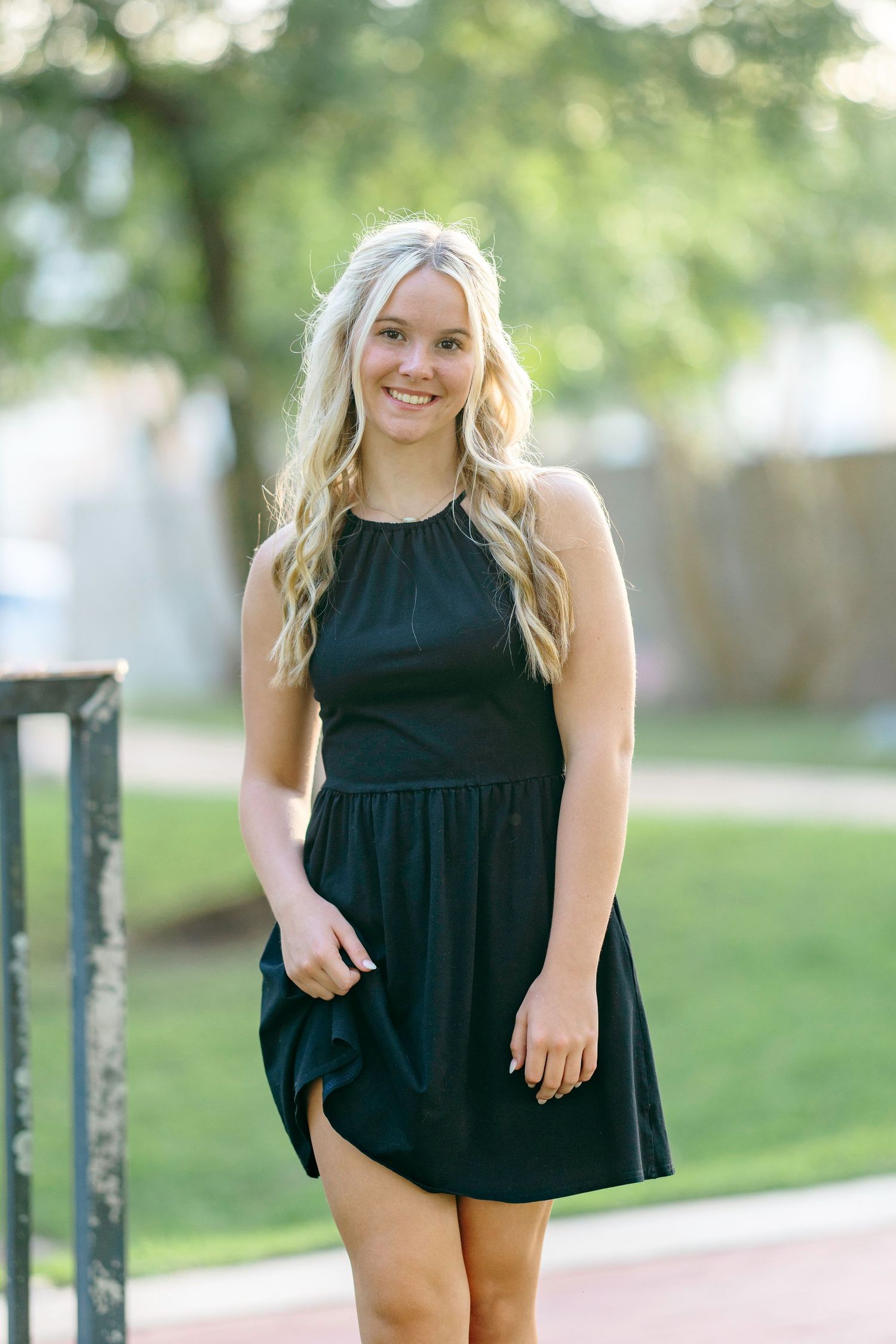 Young blonde woman smiling in a black sleeveless mini dress, posing outdoors in a park setting.