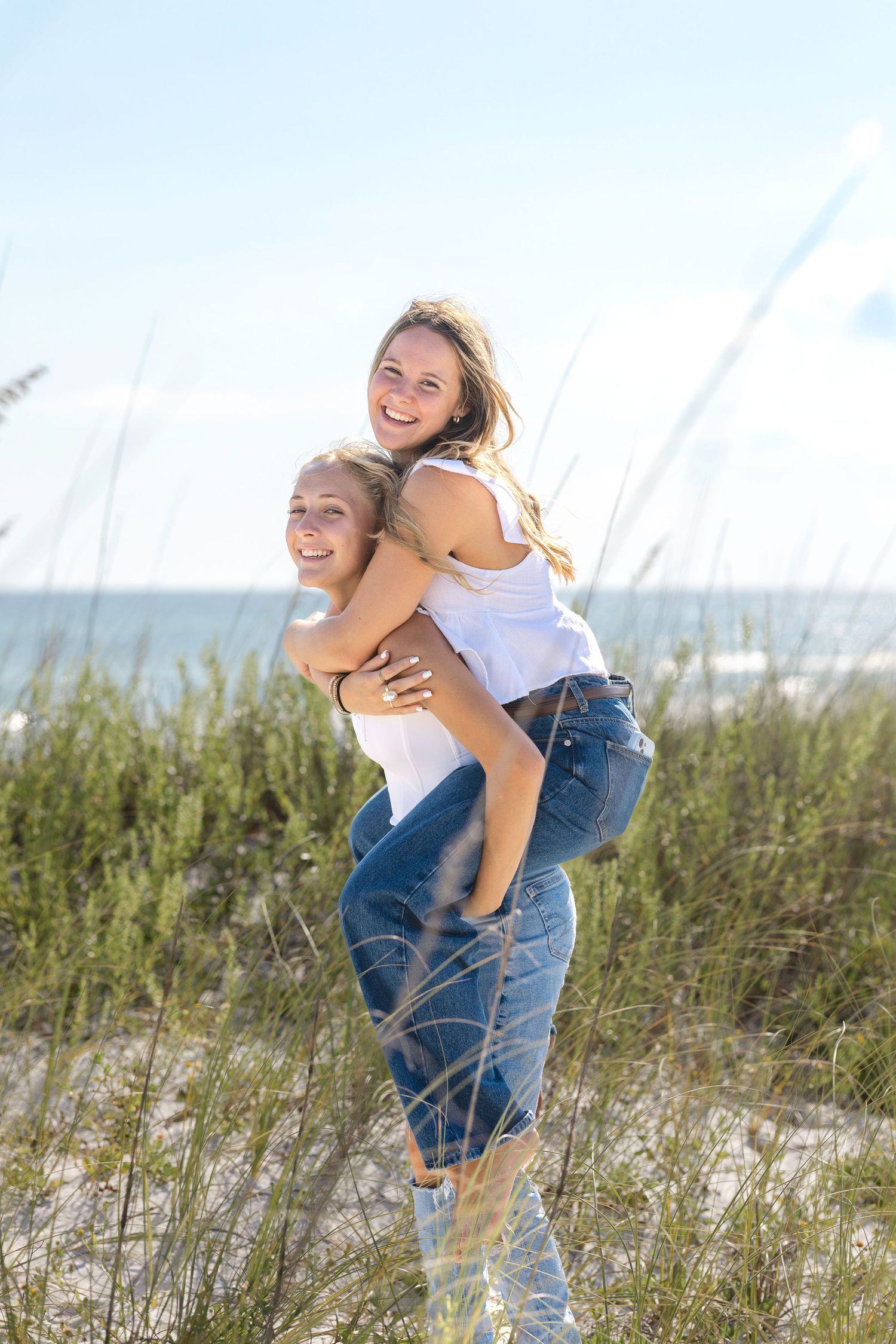 A playful piggyback ride on the beach with sand dunes and ocean in the background on a sunny day.