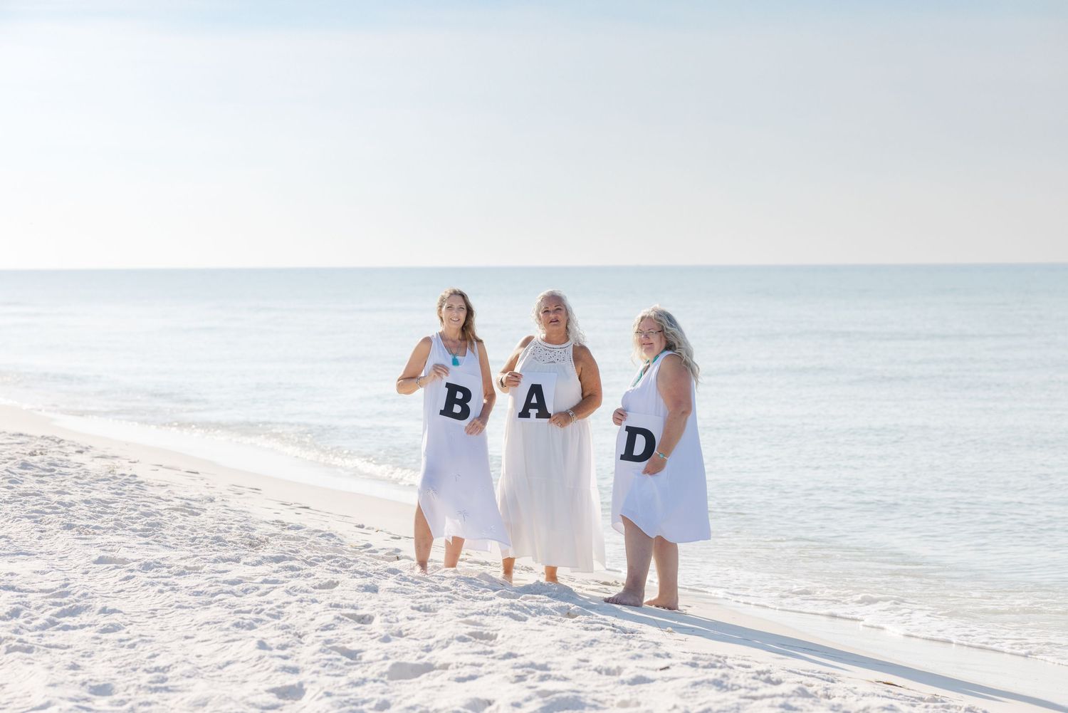 Three people in white dresses wearing navy blue letters spelling DAD walk together on a sunny beach by the ocean.