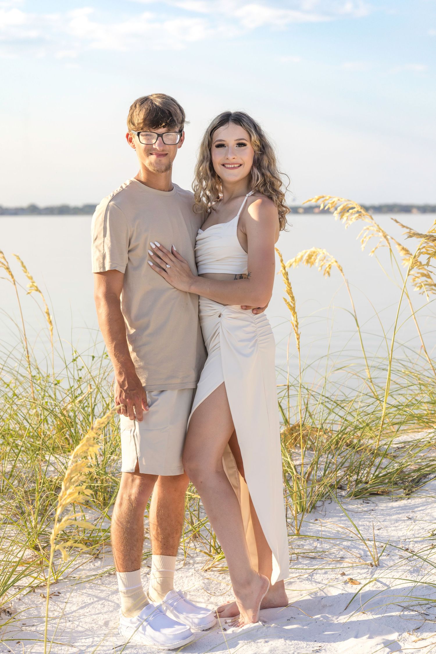 A couple poses together on a sandy beach at sunset with sea oats and water in the background.