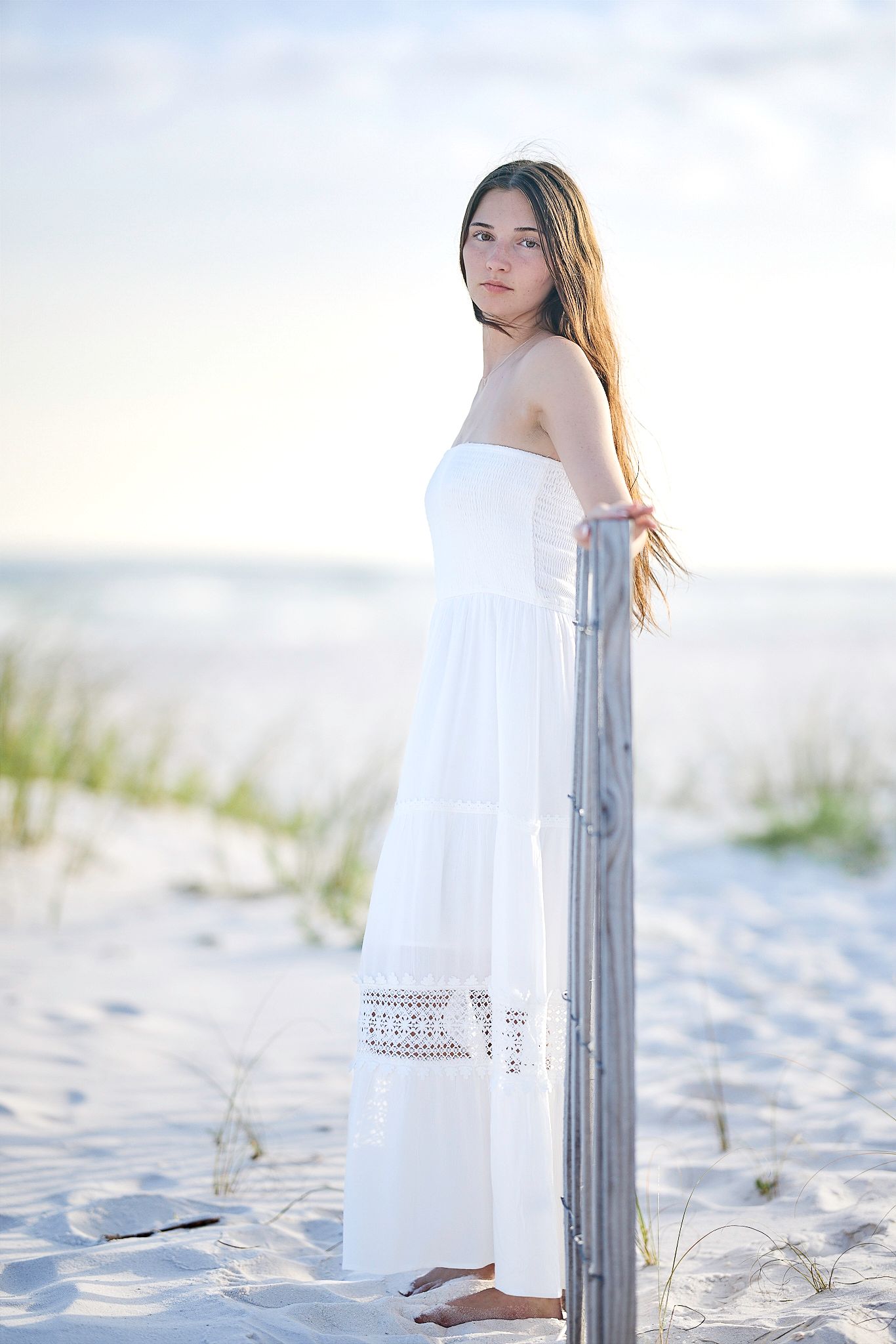 A person in a flowing white dress stands on a sandy beach next to a wooden post at sunset.