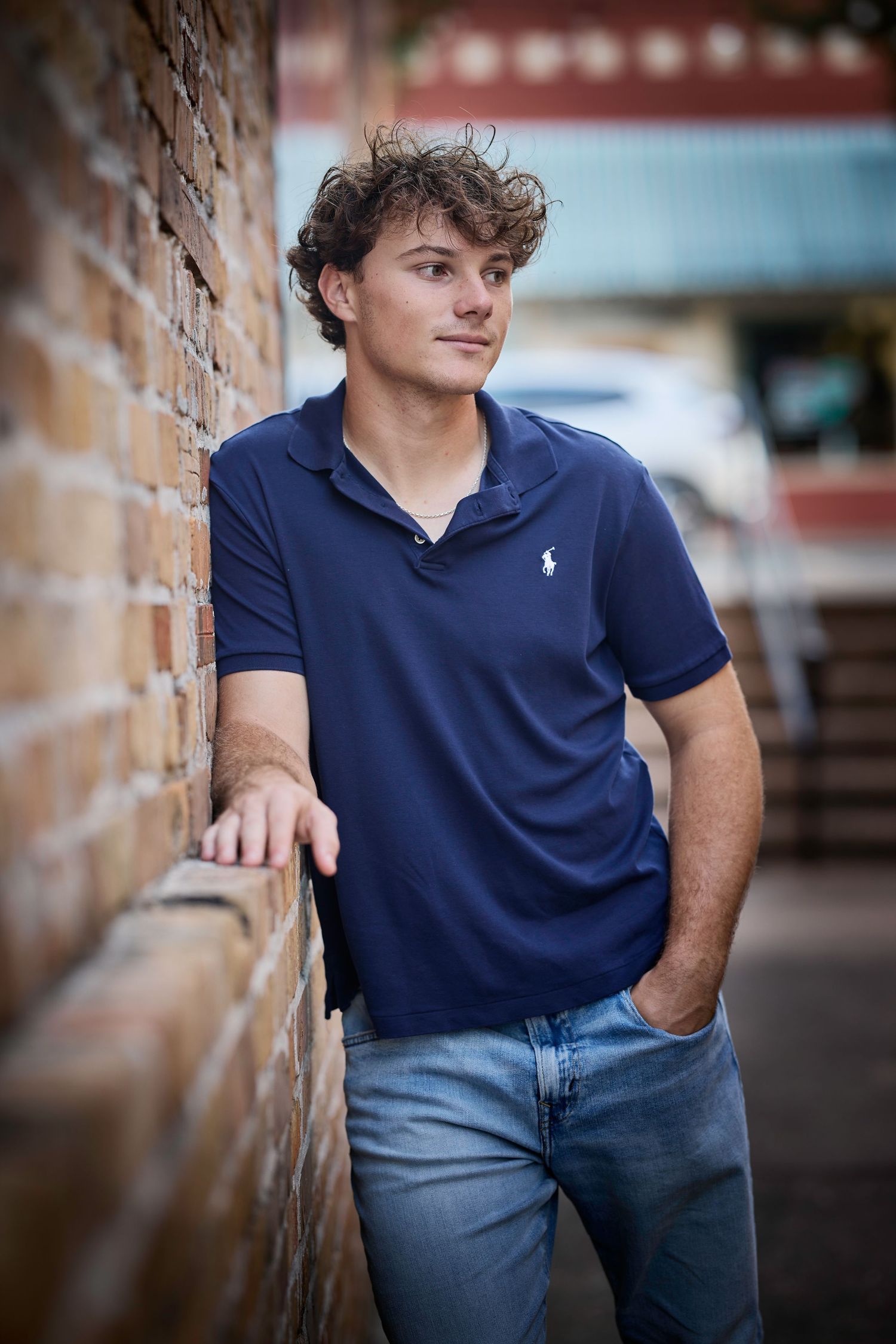 Person in navy polo shirt and jeans poses casually against brick wall in outdoor portrait session.