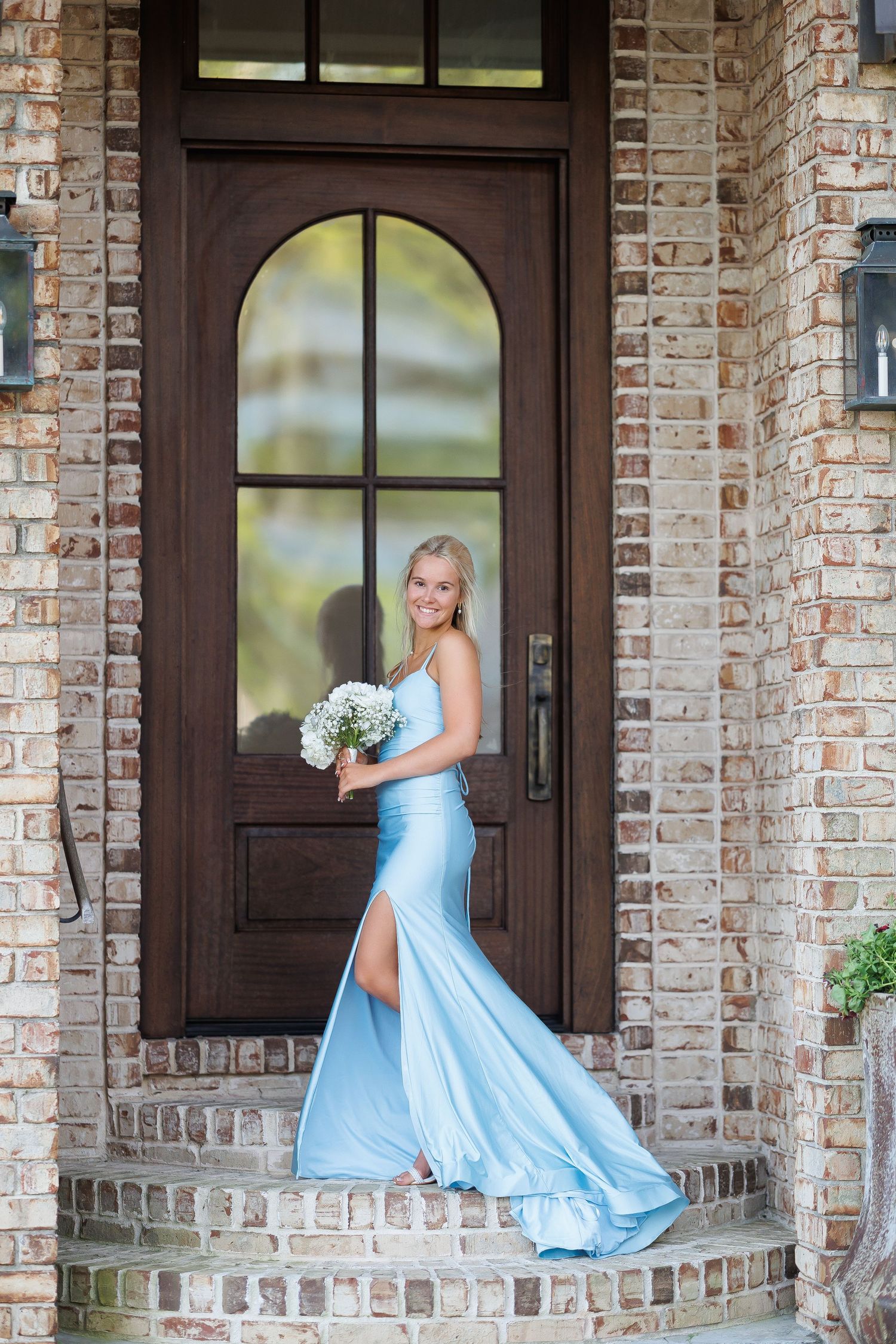 An elegant light blue formal gown with a bouquet stands against a dark wooden door and brick exterior.