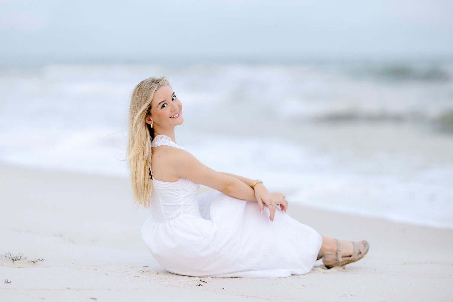 A woman in a flowing white dress sits peacefully on a sandy beach with ocean waves in the background.