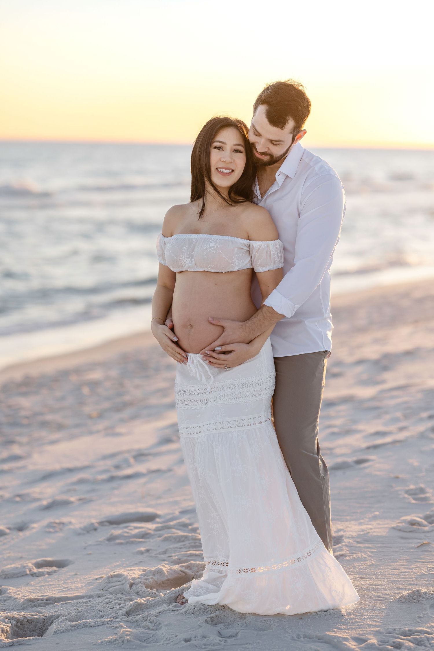A couple embraces on a sandy beach at sunset, with the woman wearing a white lace dress and the man in casual attire.