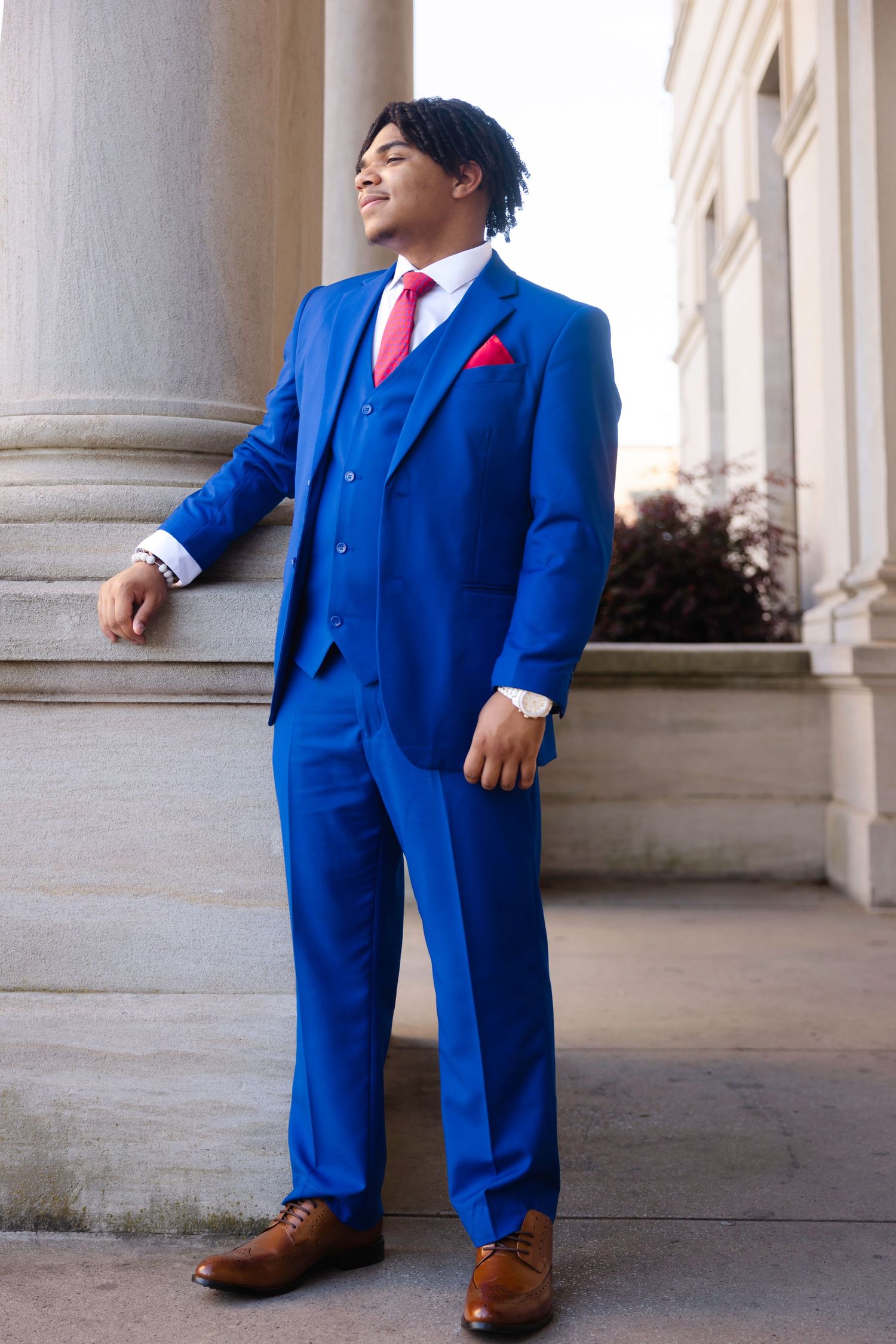 Young man in a royal blue three-piece suit with red tie and pocket square, posing by columns.