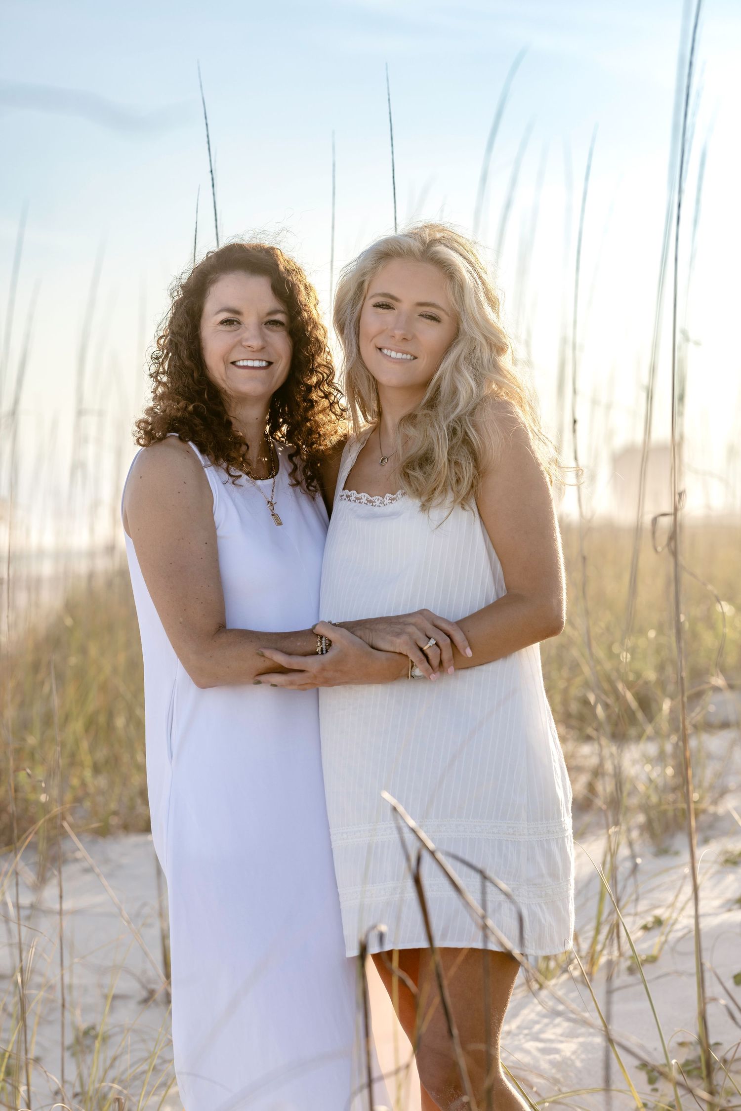 Two smiling women in white dresses pose together on a sandy beach with sea oats at golden hour.