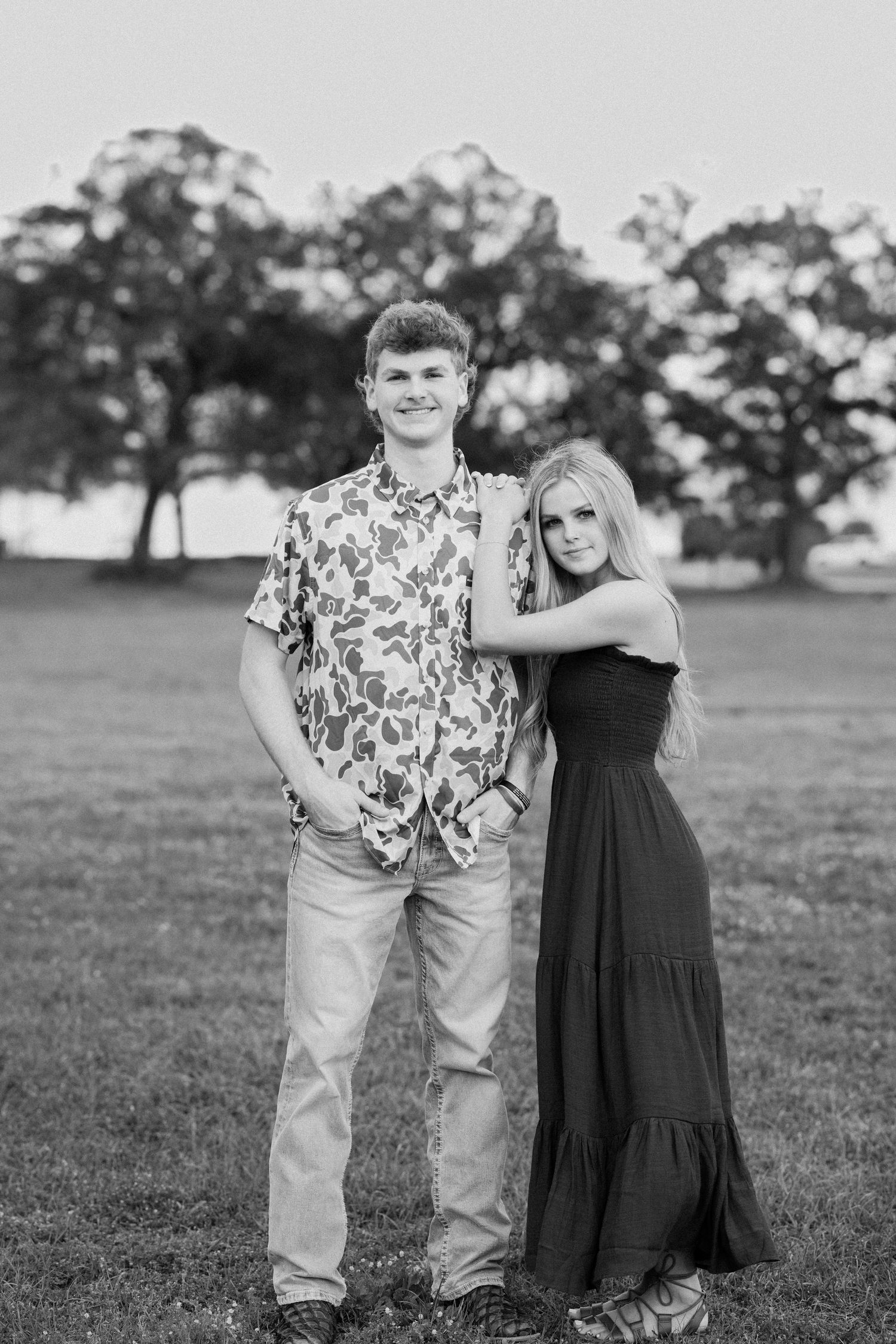 A couple poses together in a grassy field wearing contrasting floral print and black dress outfits.