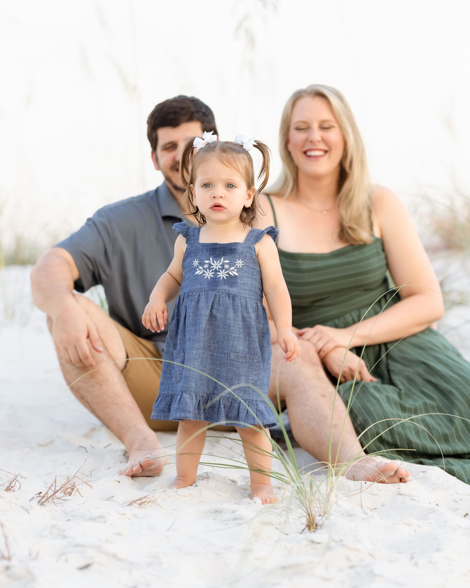 Family of three poses on white sand beach, parents sitting while toddler girl in blue dress stands between them.