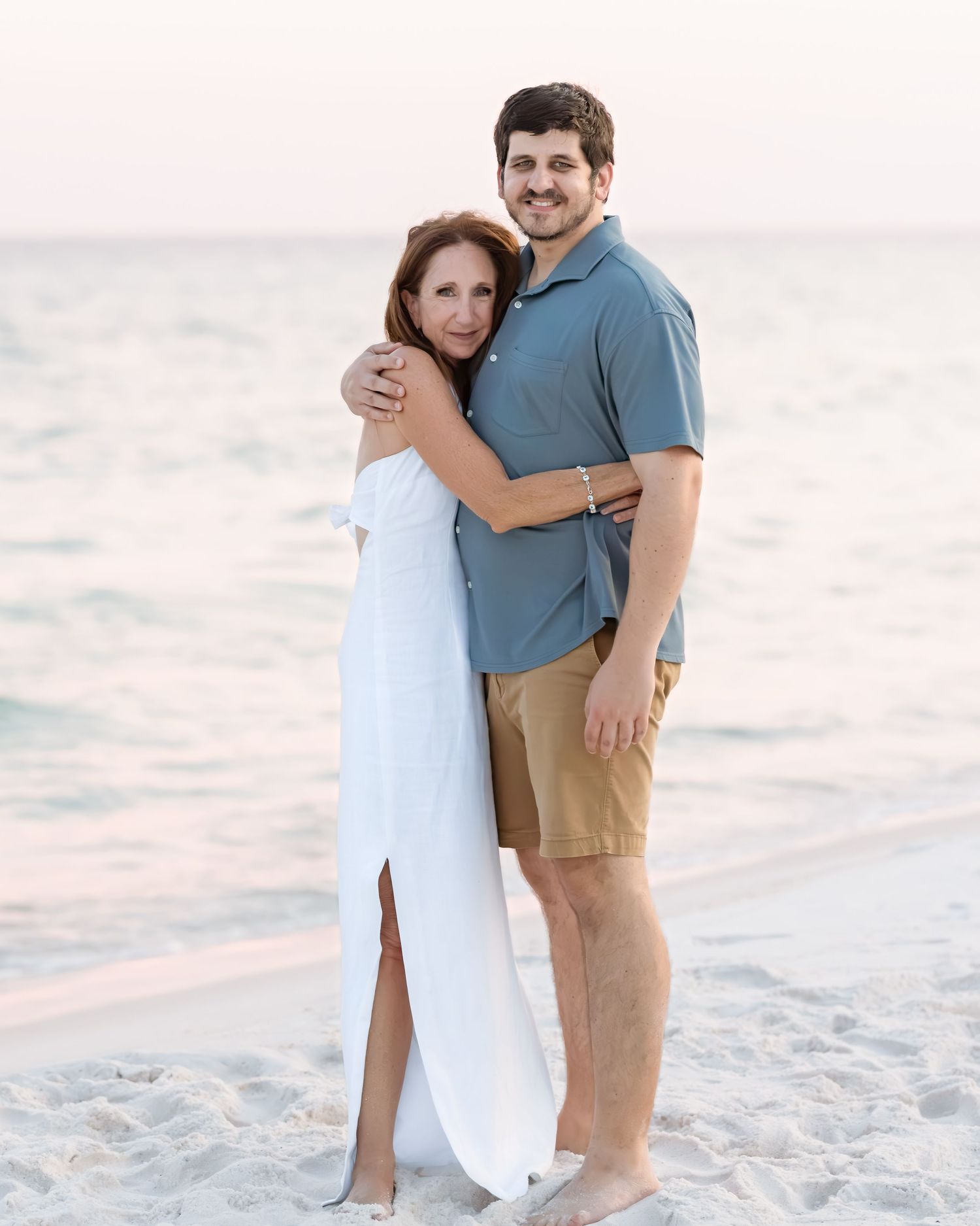 Couple hugging on white sand beach at sunset, woman in white dress, man in blue polo and khaki shorts.