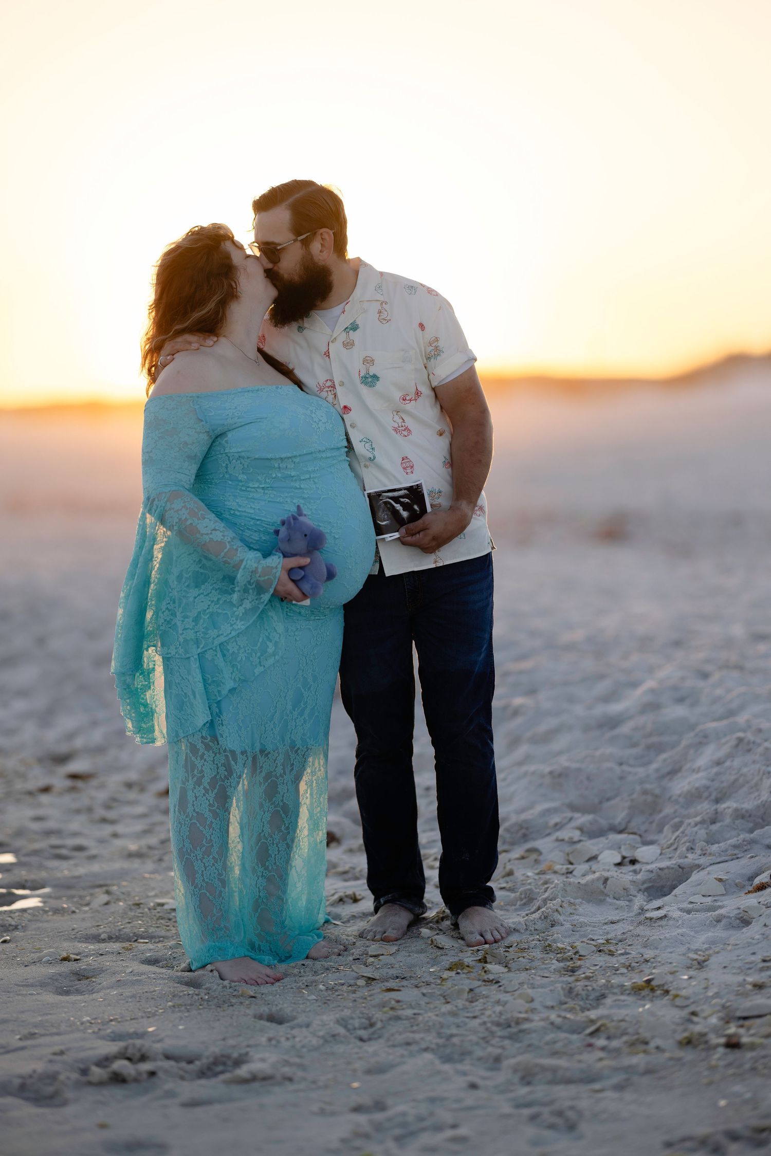 Pregnant couple kissing on beach at sunset, woman in teal lace gown holding flowers, man holding ultrasound photo.