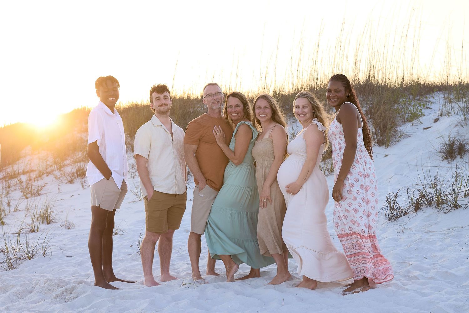 Group of seven adults posing on a sandy beach at golden sunset with sand dunes behind them.