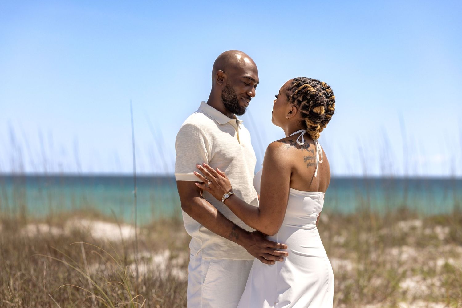 A couple in white attire embraces on a sandy beach with ocean waves and sea grass in the background.