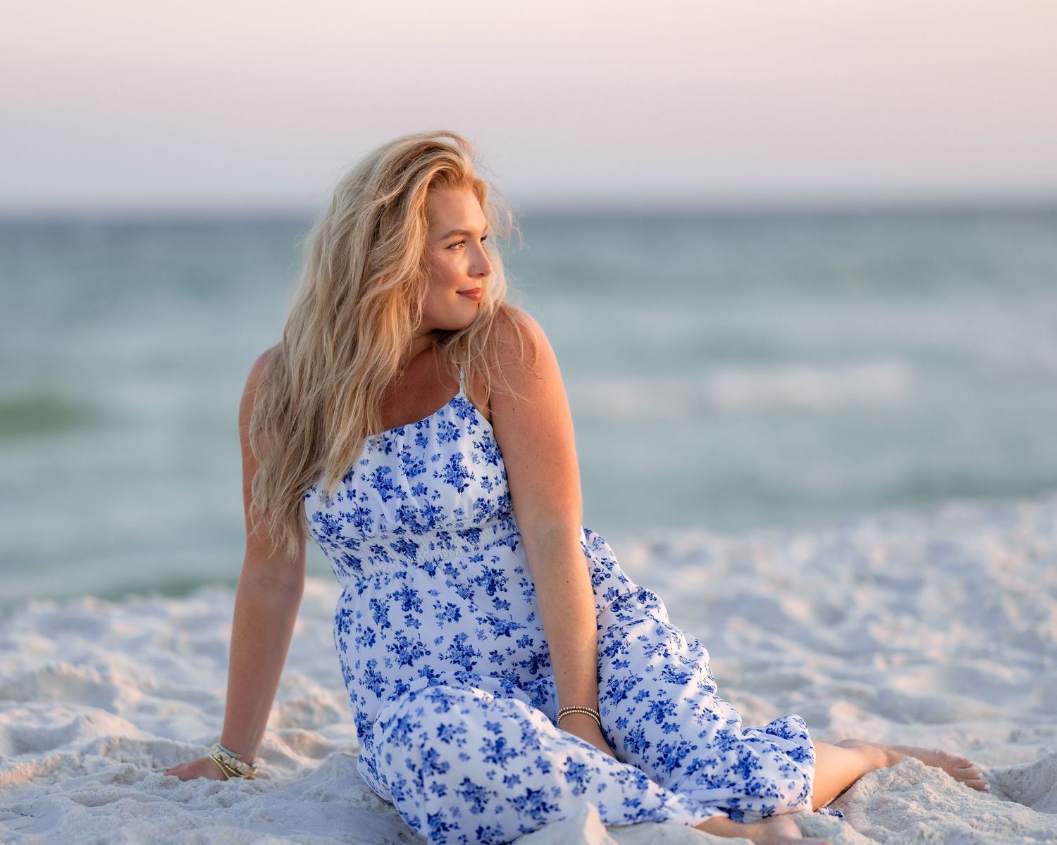A blonde woman in a blue floral dress sits gazing at the beach horizon.