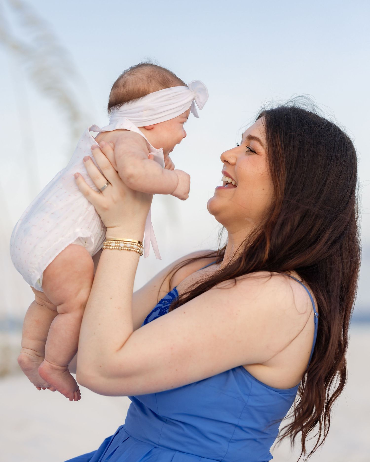 Joyful young mother in blue dress lifting and smiling at her baby on a sunny beach.