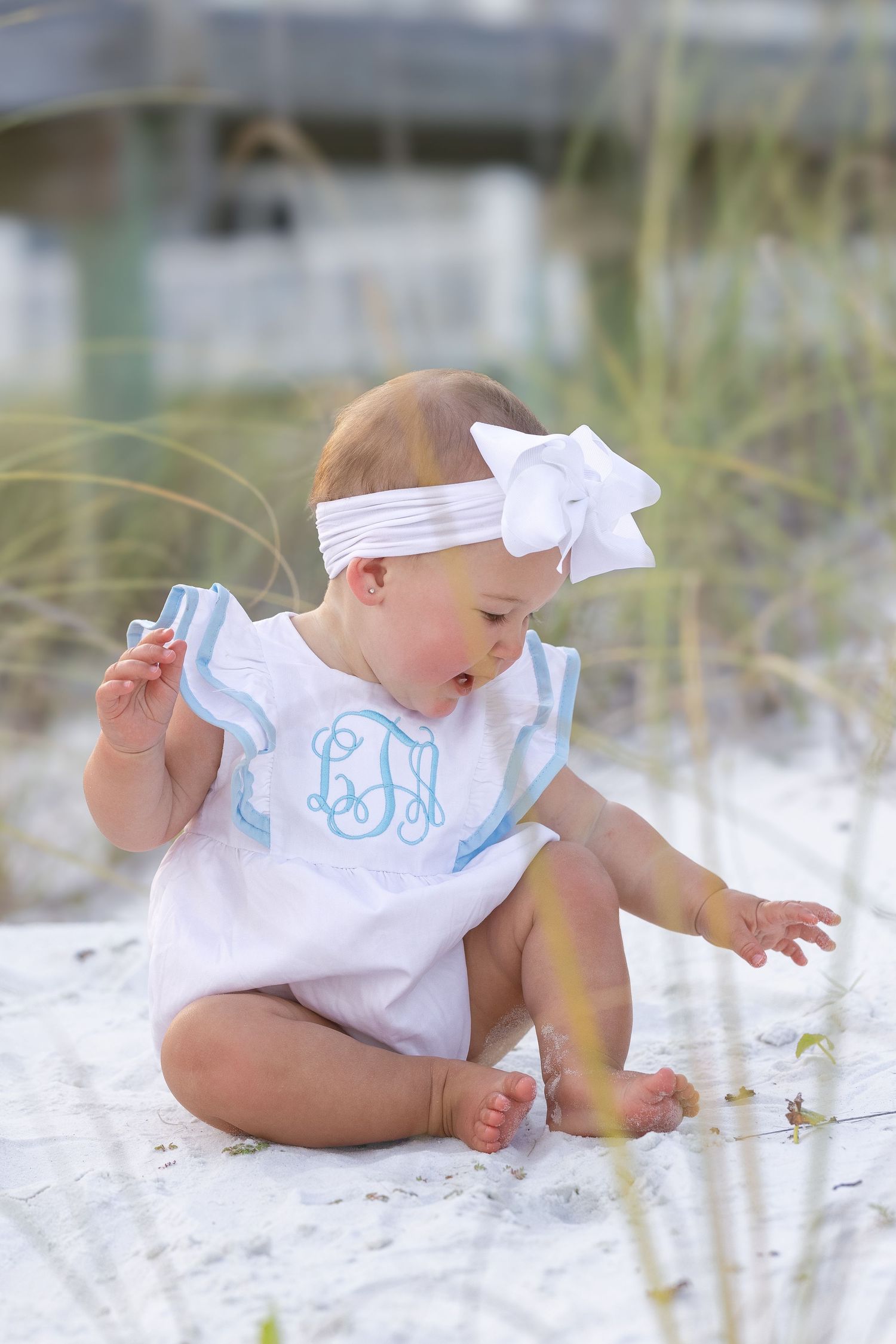 Baby girl in white monogrammed romper and bow headband sitting on white sand beach.