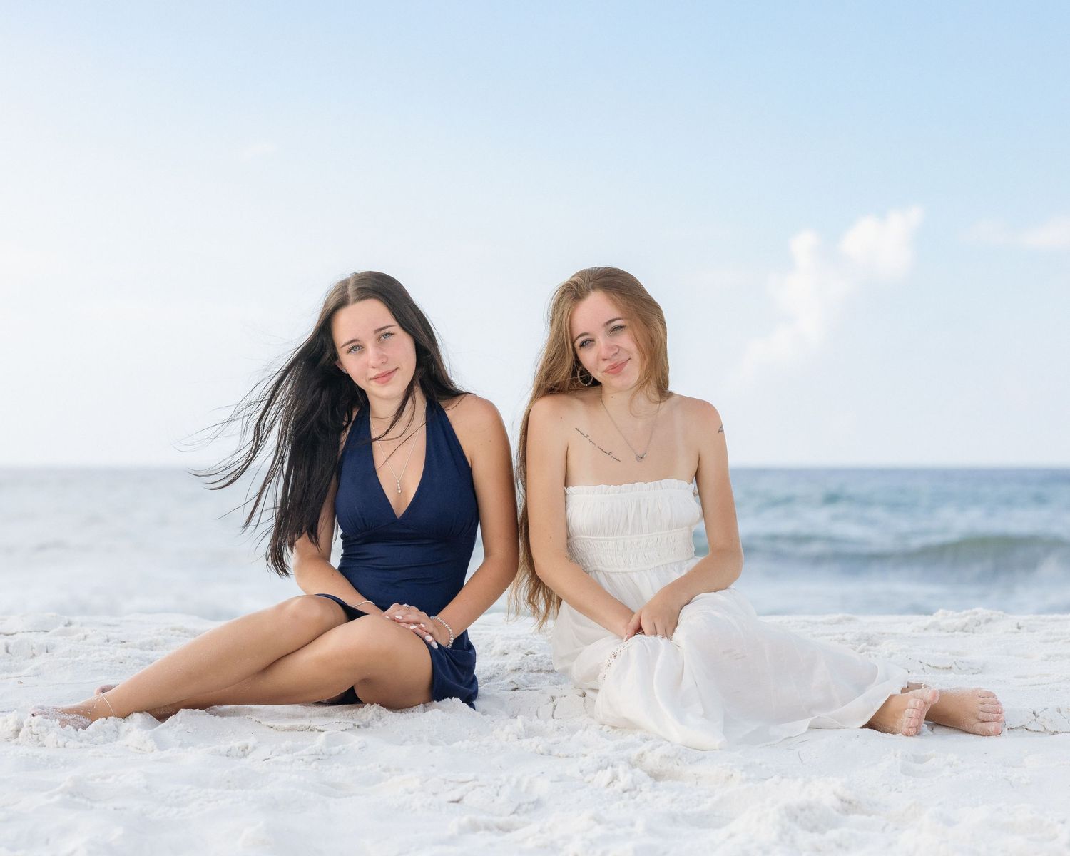 Two young women sitting on white sand beach, one in navy swimsuit, one in white dress, ocean backdrop.