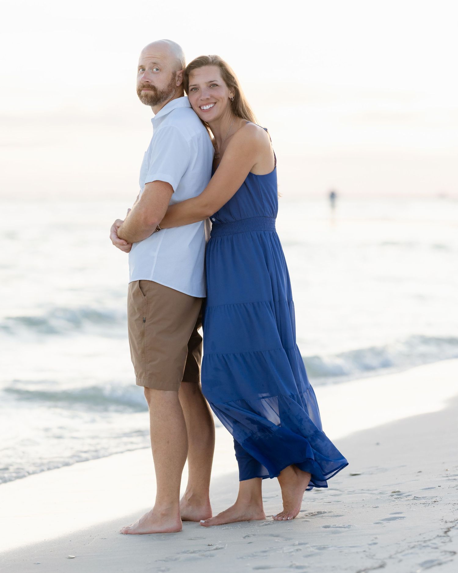 A smiling couple poses back-to-back on a beach at sunset, woman in blue maxi dress, man in khaki shorts.