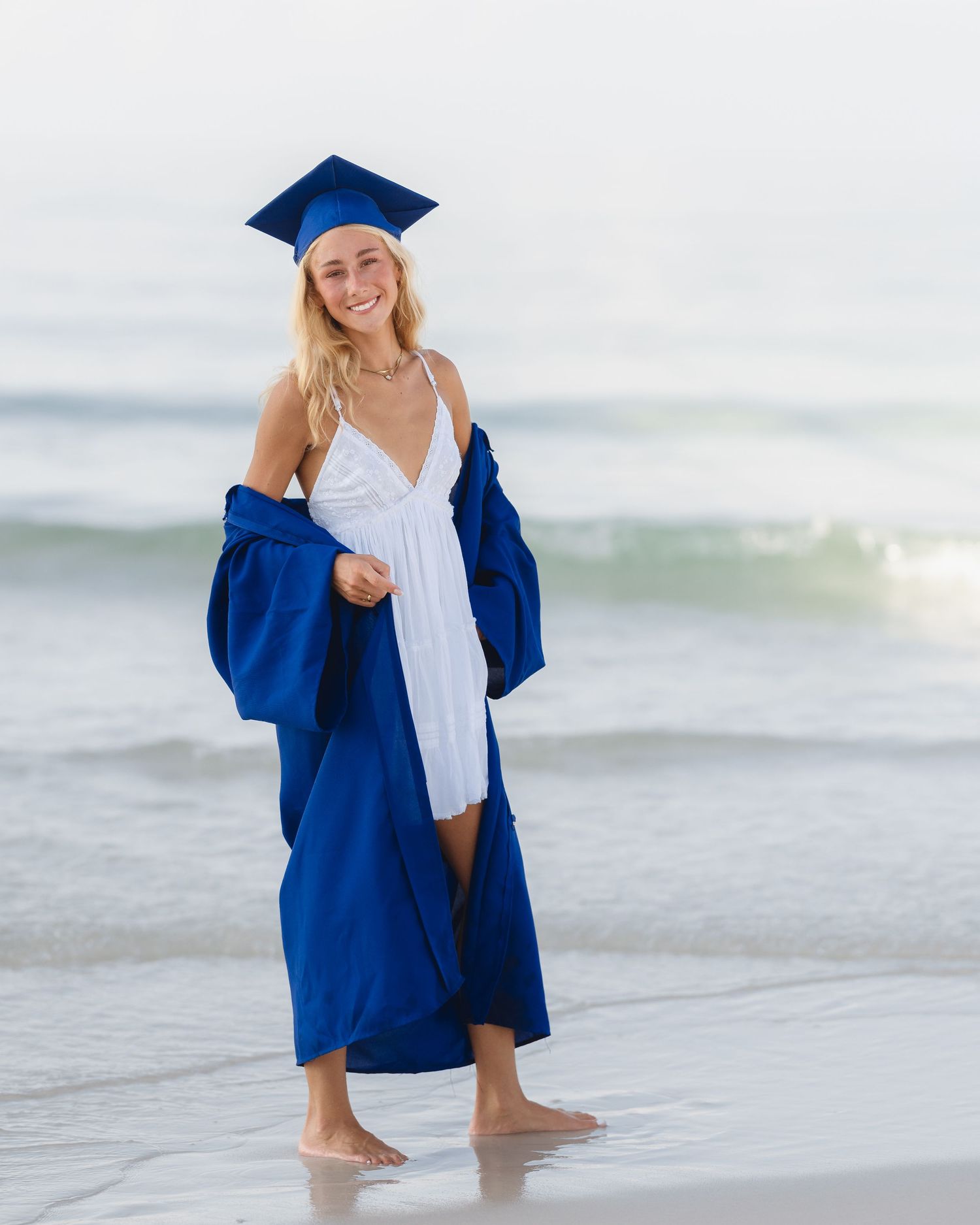 Smiling graduate in blue cap and gown over white dress standing barefoot on a sandy beach with ocean waves.