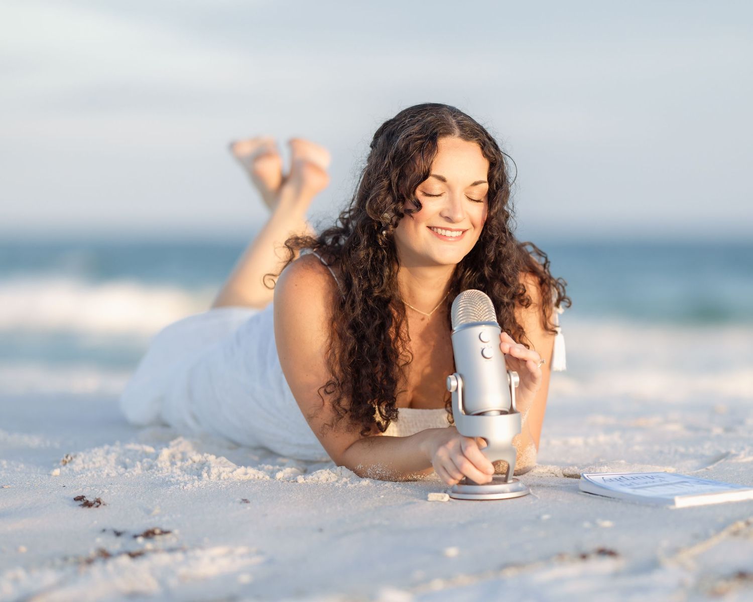 Young woman lying on beach sand smiling at blue podcast microphone on sunny day.