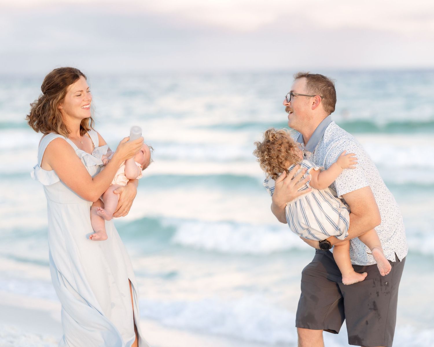 Family of four enjoying a beach day, parents holding two young children with ocean waves in the background.