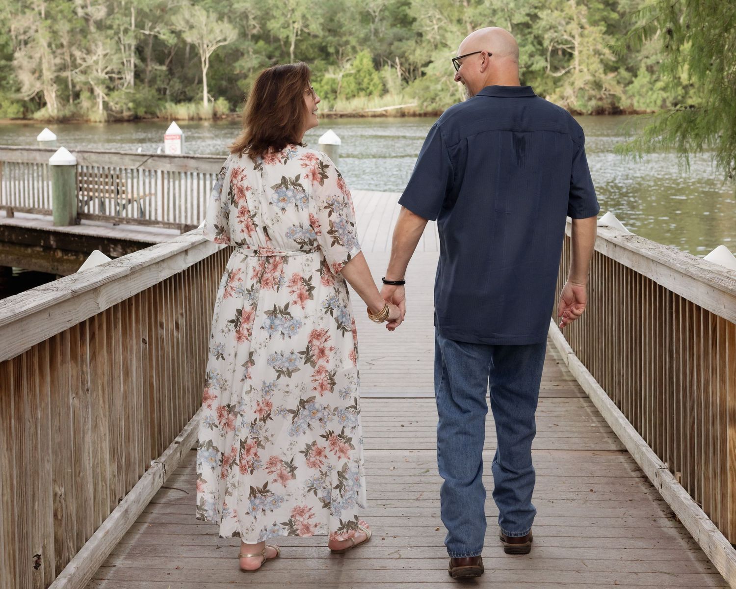 Couple holding hands walking away on a wooden bridge over a scenic lake surrounded by trees.