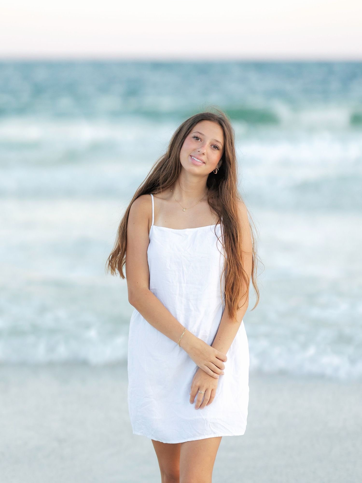 Young woman in white sundress standing on beach with ocean waves in background.
