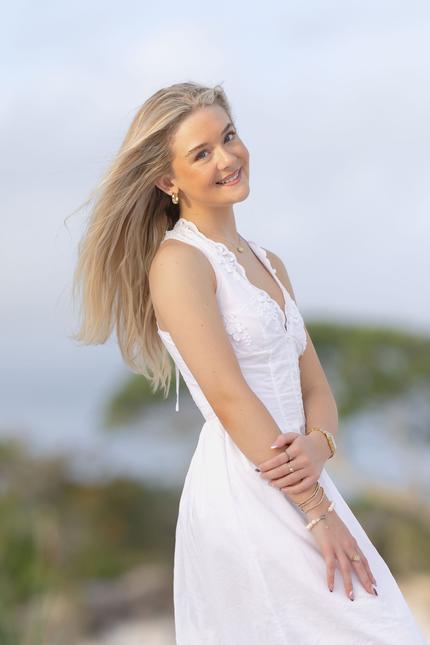 Smiling blonde woman in white sleeveless dress with gold bracelets, hair blowing in wind outdoors.