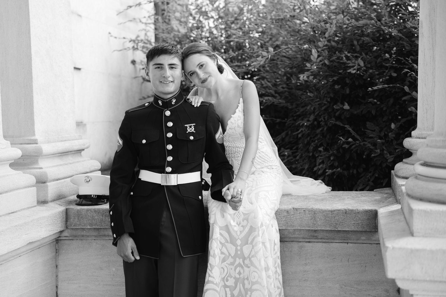 Black and white wedding photo of a Marine in dress uniform and bride in lace gown posing on stone steps.