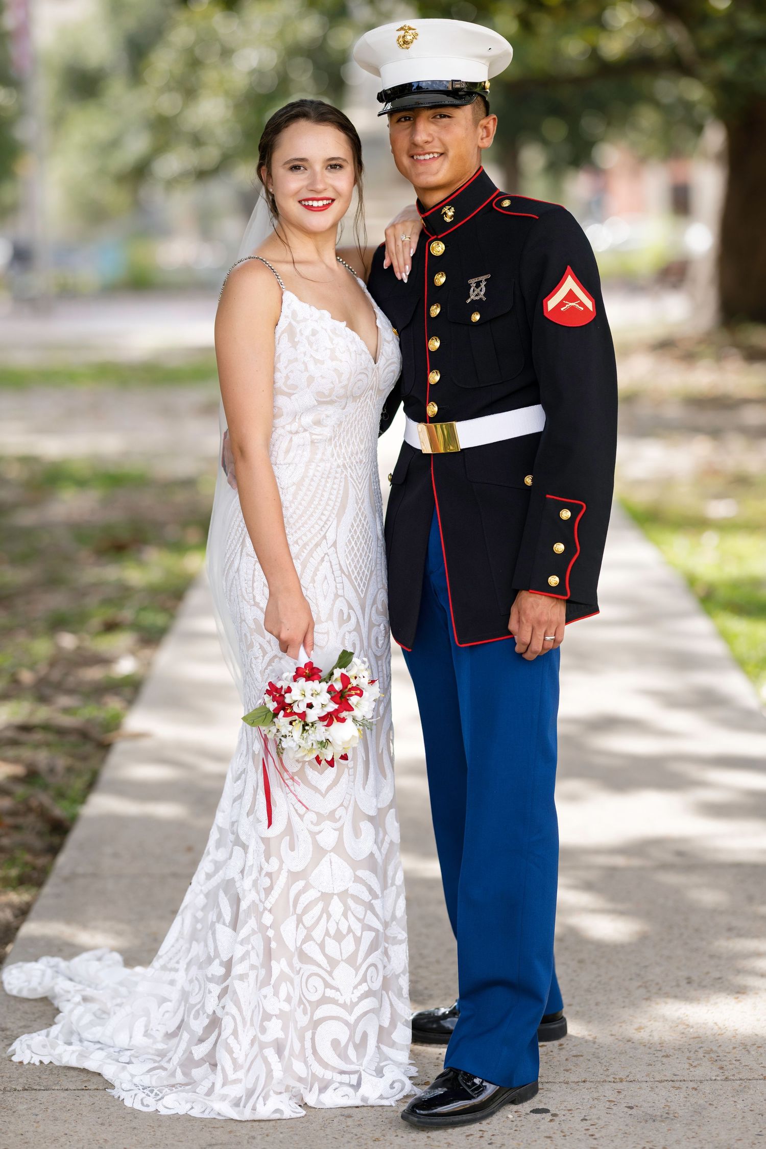 A bride in white lace gown and groom in Marine dress blues uniform pose together outdoors on their wedding day.