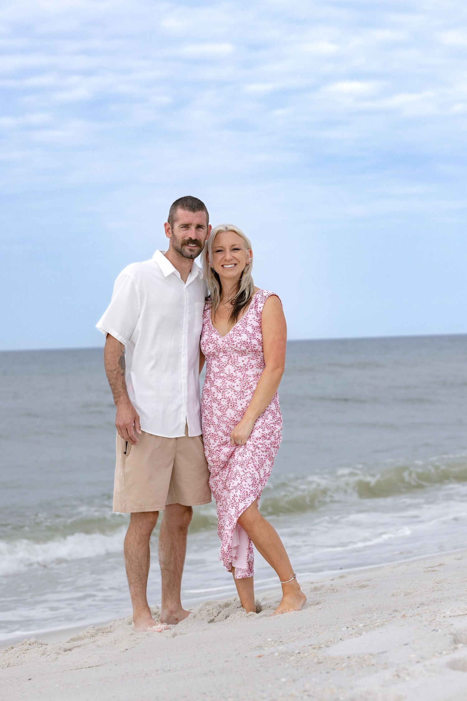 A smiling couple stands together on a sandy beach with ocean waves behind them on a cloudy day.