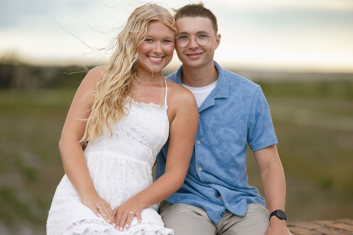 A smiling young couple poses outdoors at sunset, woman in white dress and man in blue shirt, with green fields behind.