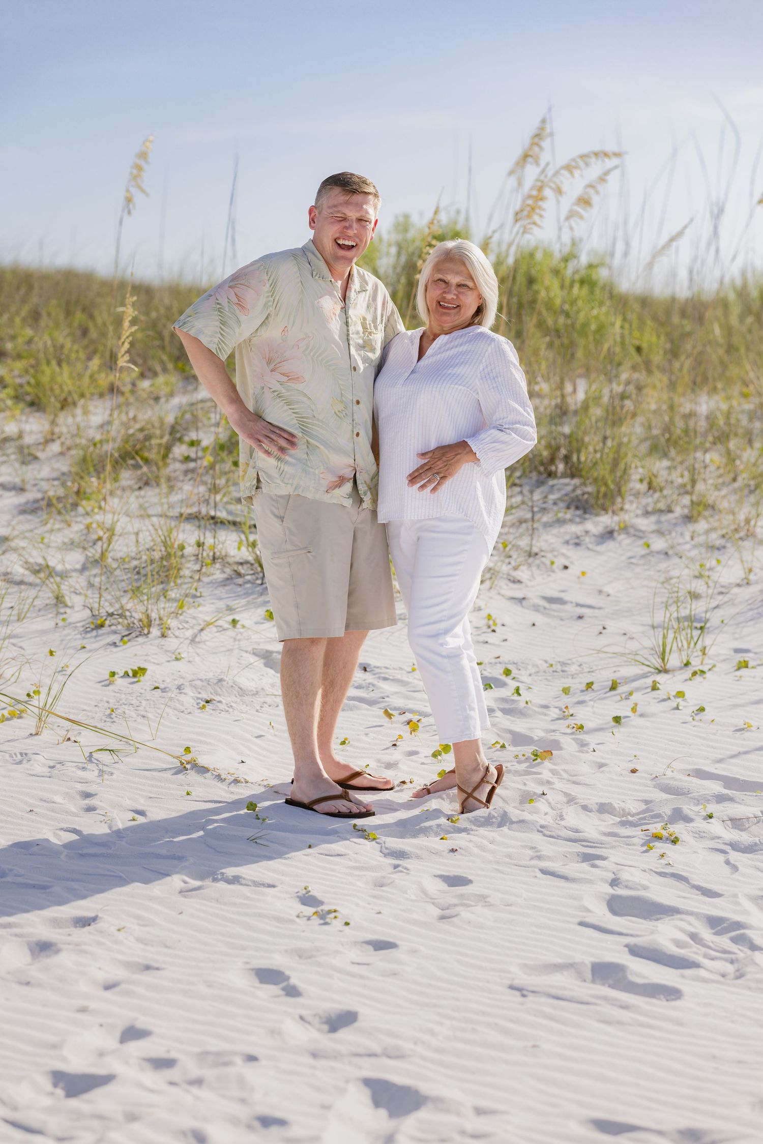 A smiling couple poses together on white sand beach dunes with sea oats in the background on a sunny day.