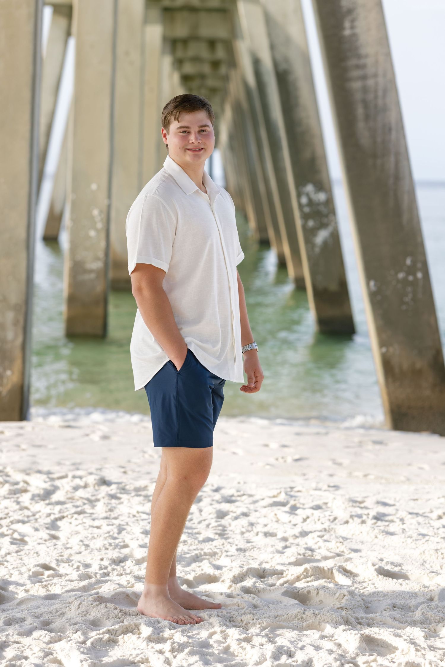 Young man in white shirt and navy shorts standing barefoot on white sand beach near a pier.