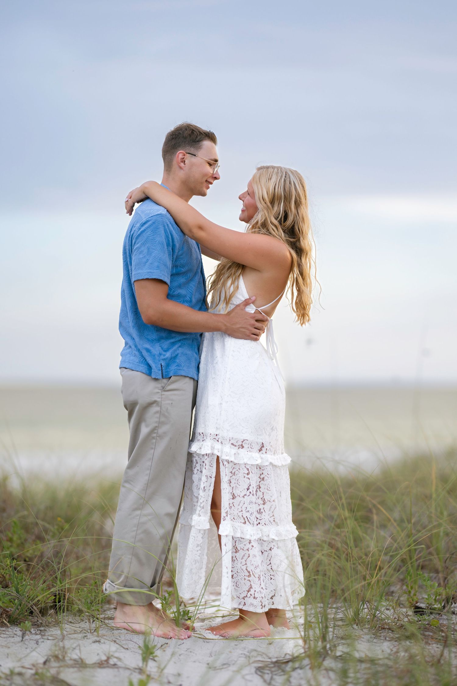 A barefoot couple embraces and gazes at each other on a sandy beach, woman in white lace dress and man in blue shirt.