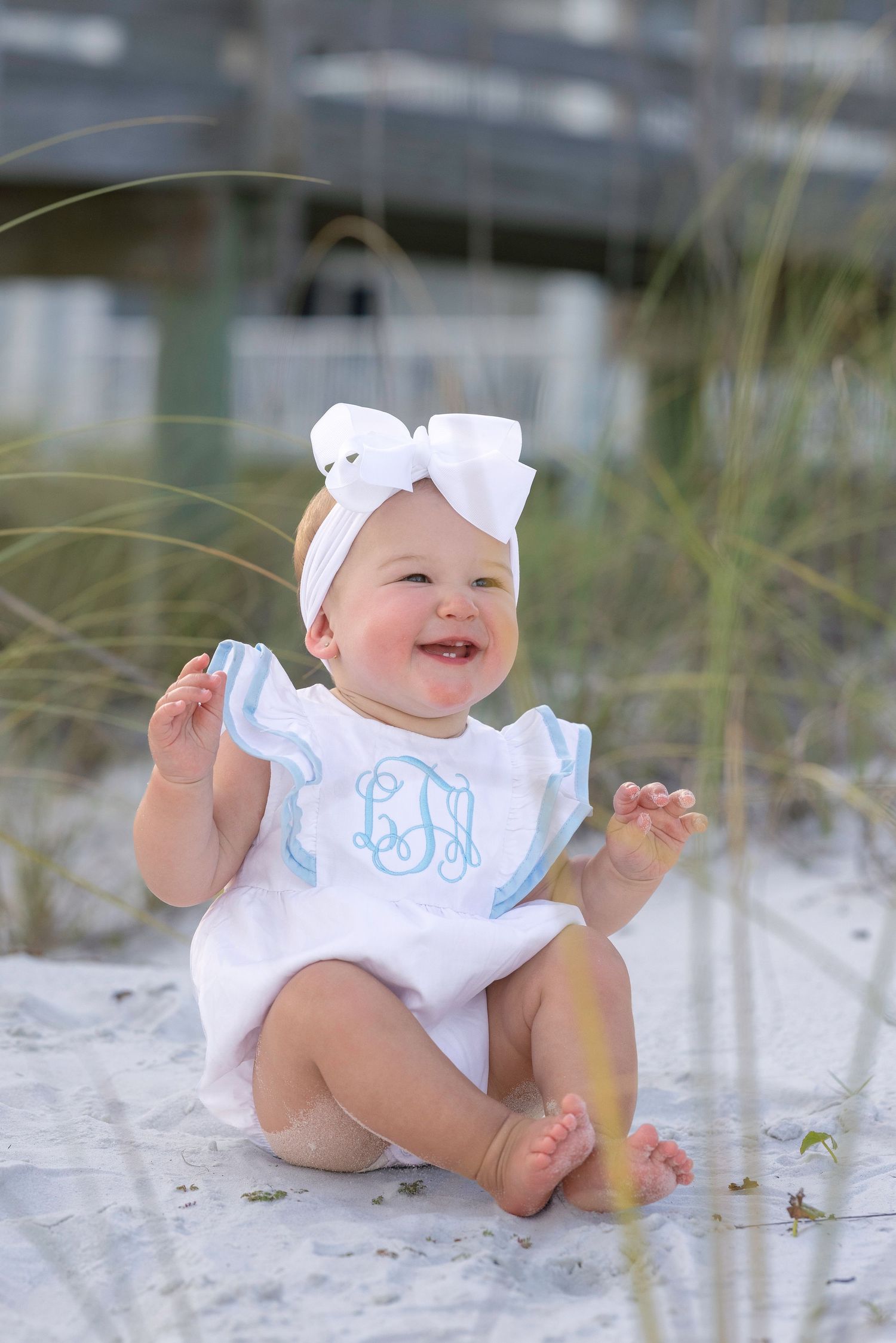 Baby girl in white monogrammed romper and bow headband smiling on white sand beach dunes.
