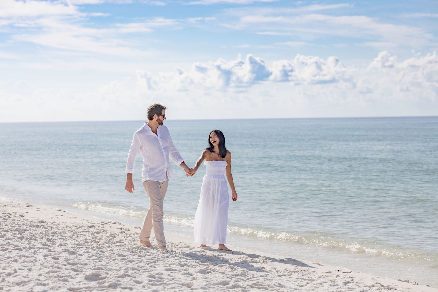 Couple in white outfits holding hands walking on a sunny white sand beach with calm turquoise ocean waters.