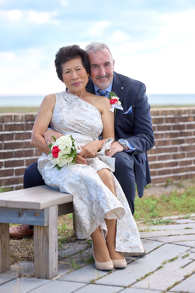A couple in wedding attire sits together on a wooden bench against a brick wall in a romantic outdoor setting.