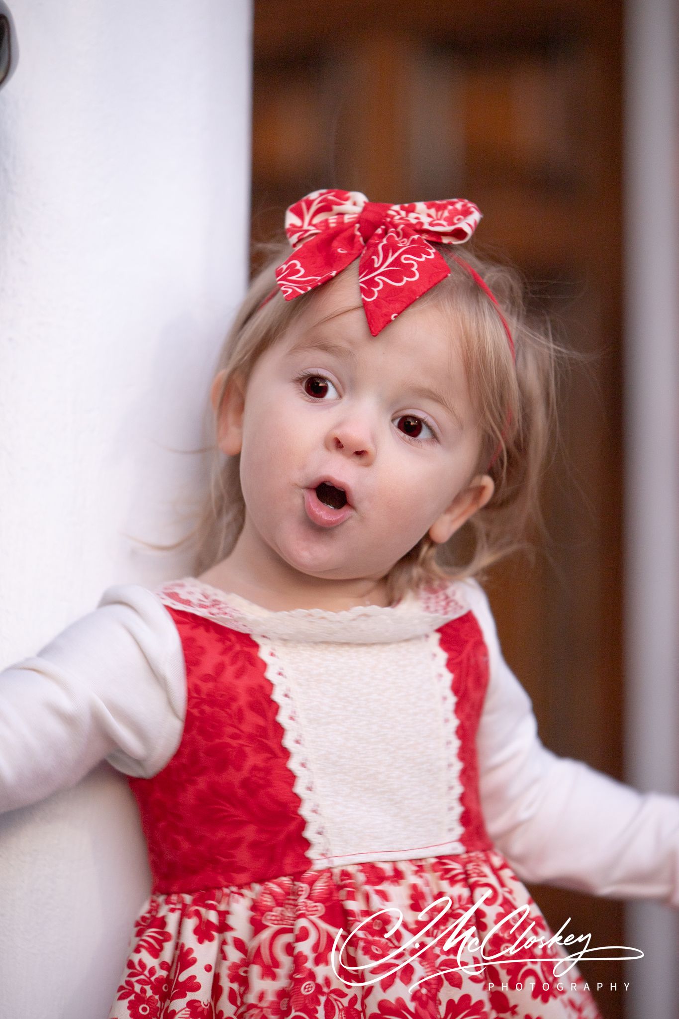 An adorable toddler in a white and red holiday dress with matching red bows makes a playful expression.