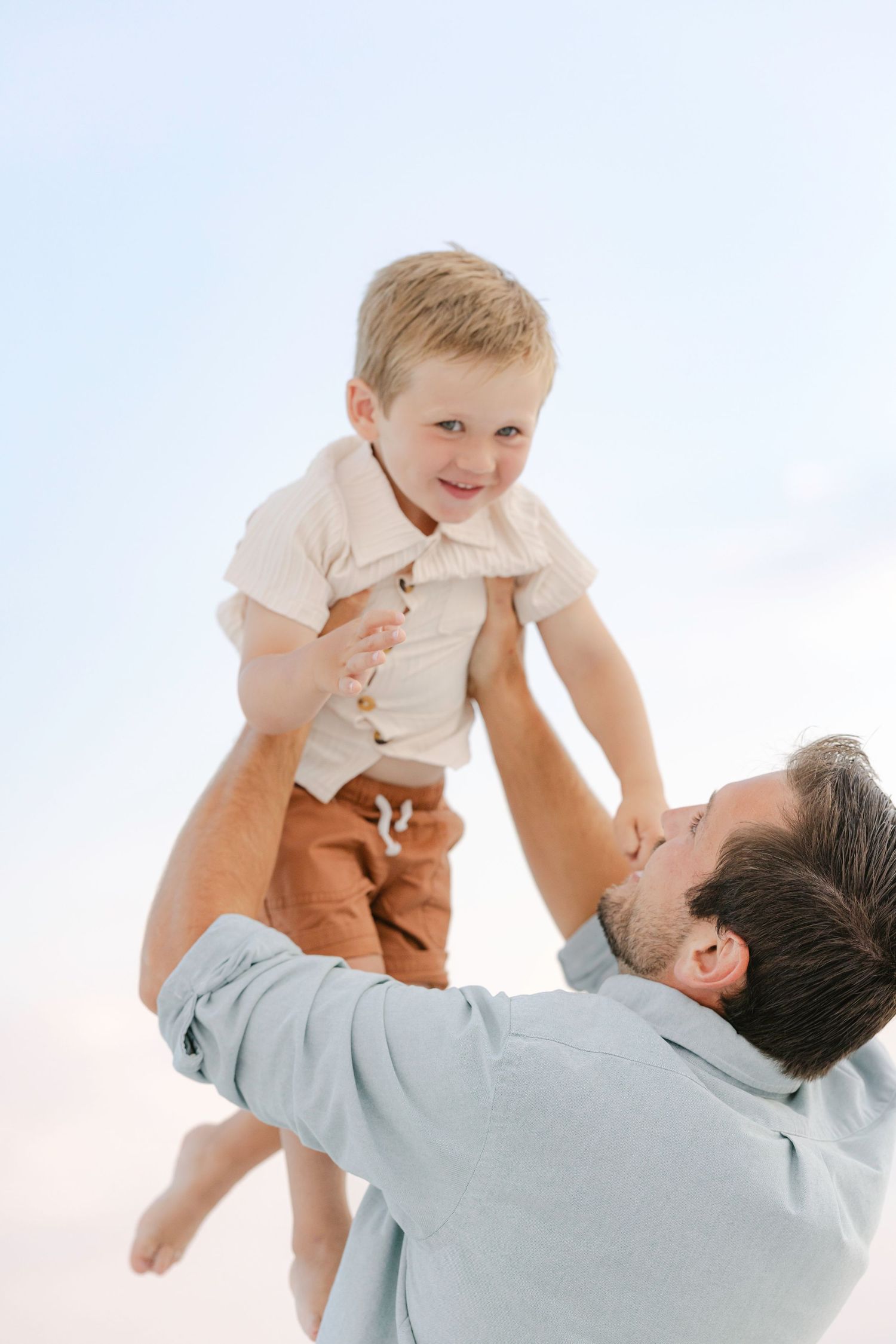 A parent wearing a light blue shirt lifts a small child wearing beige clothing playfully in the air against a white background.