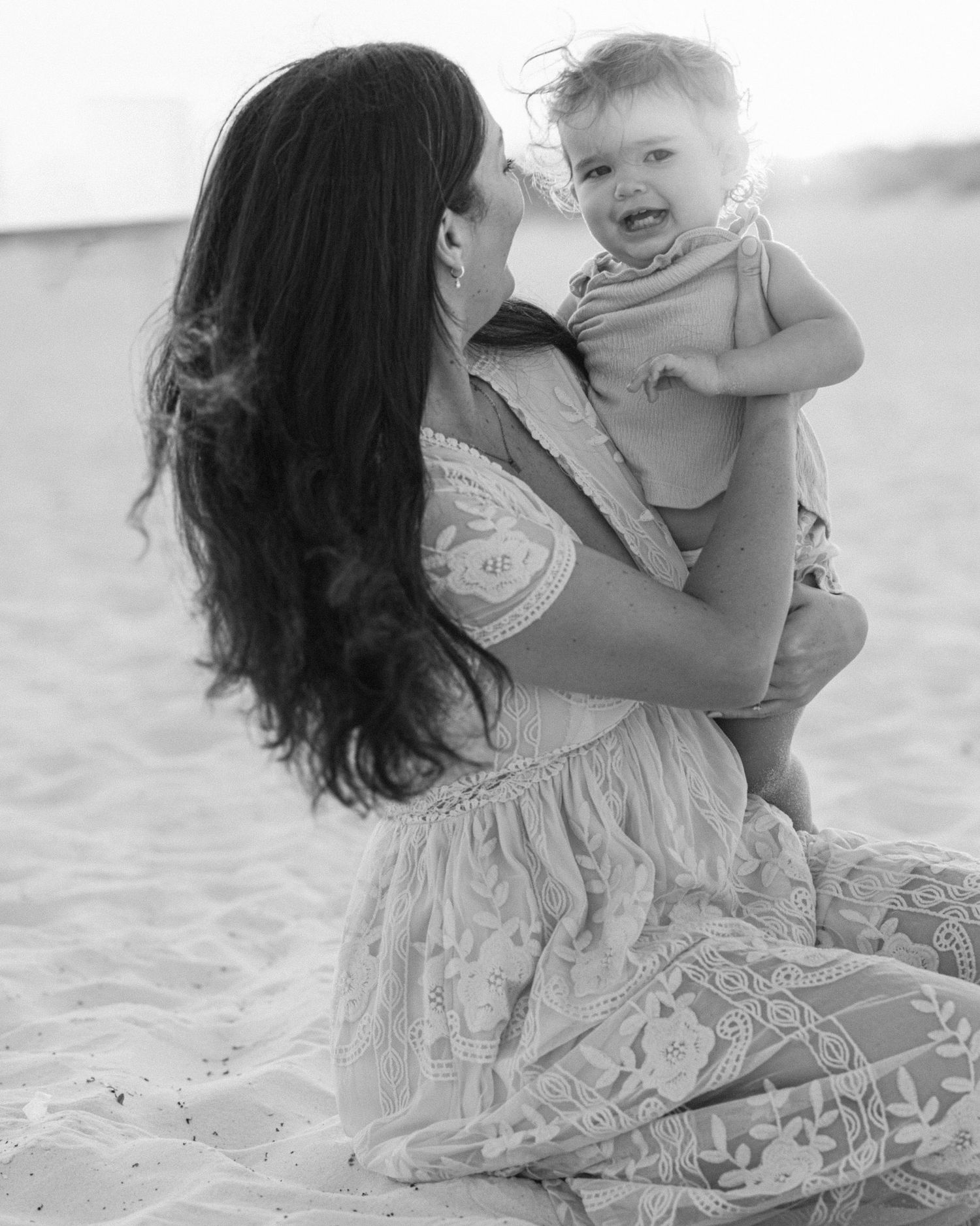 Black and white photo of a mother and baby sharing a joyful moment together on the beach.