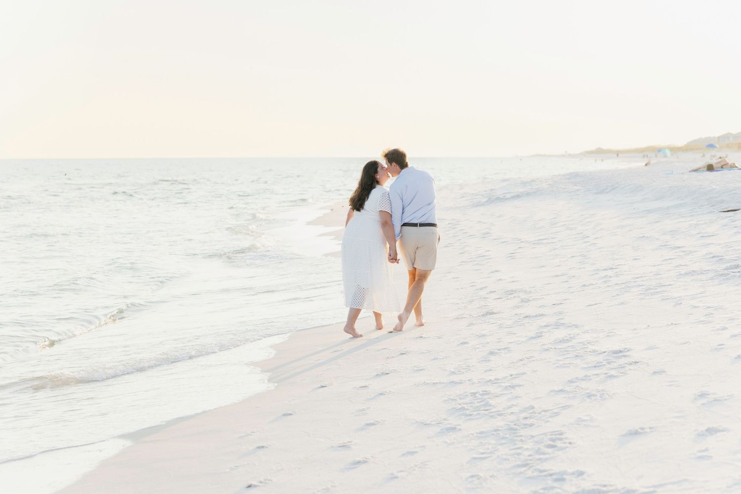 A romantic couple walks along a pristine white beach shoreline at sunset.