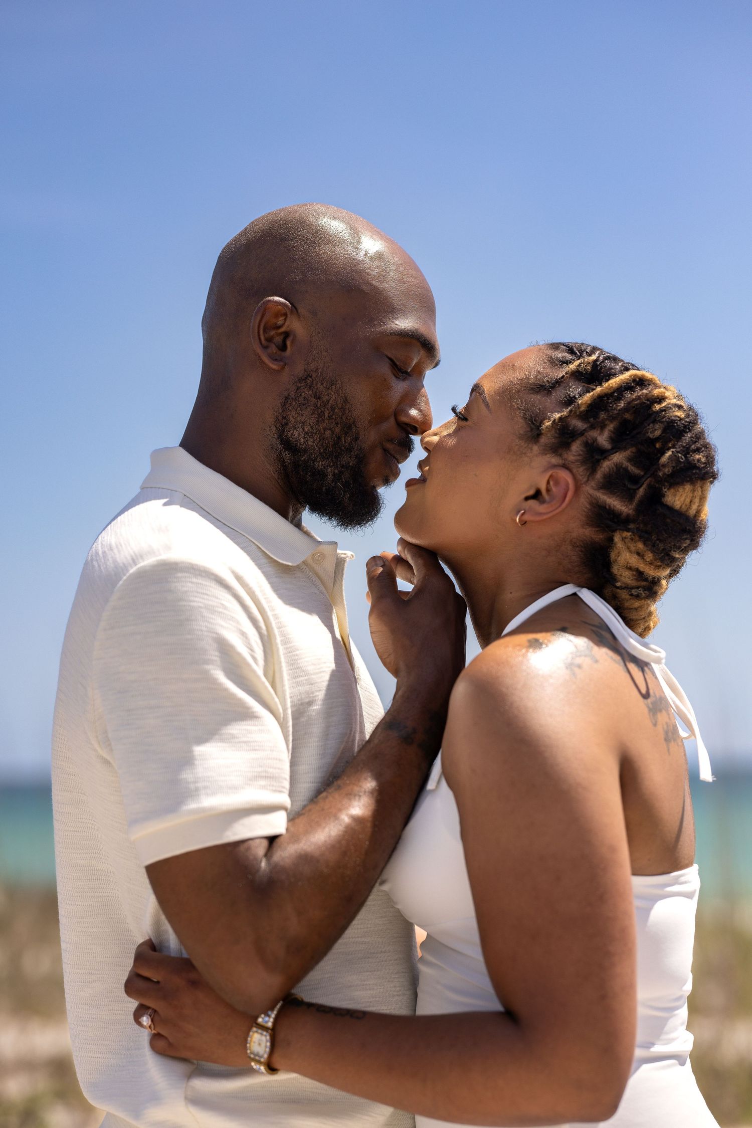 A romantic African American couple shares a tender kiss on a sunny beach, dressed in matching white outfits.