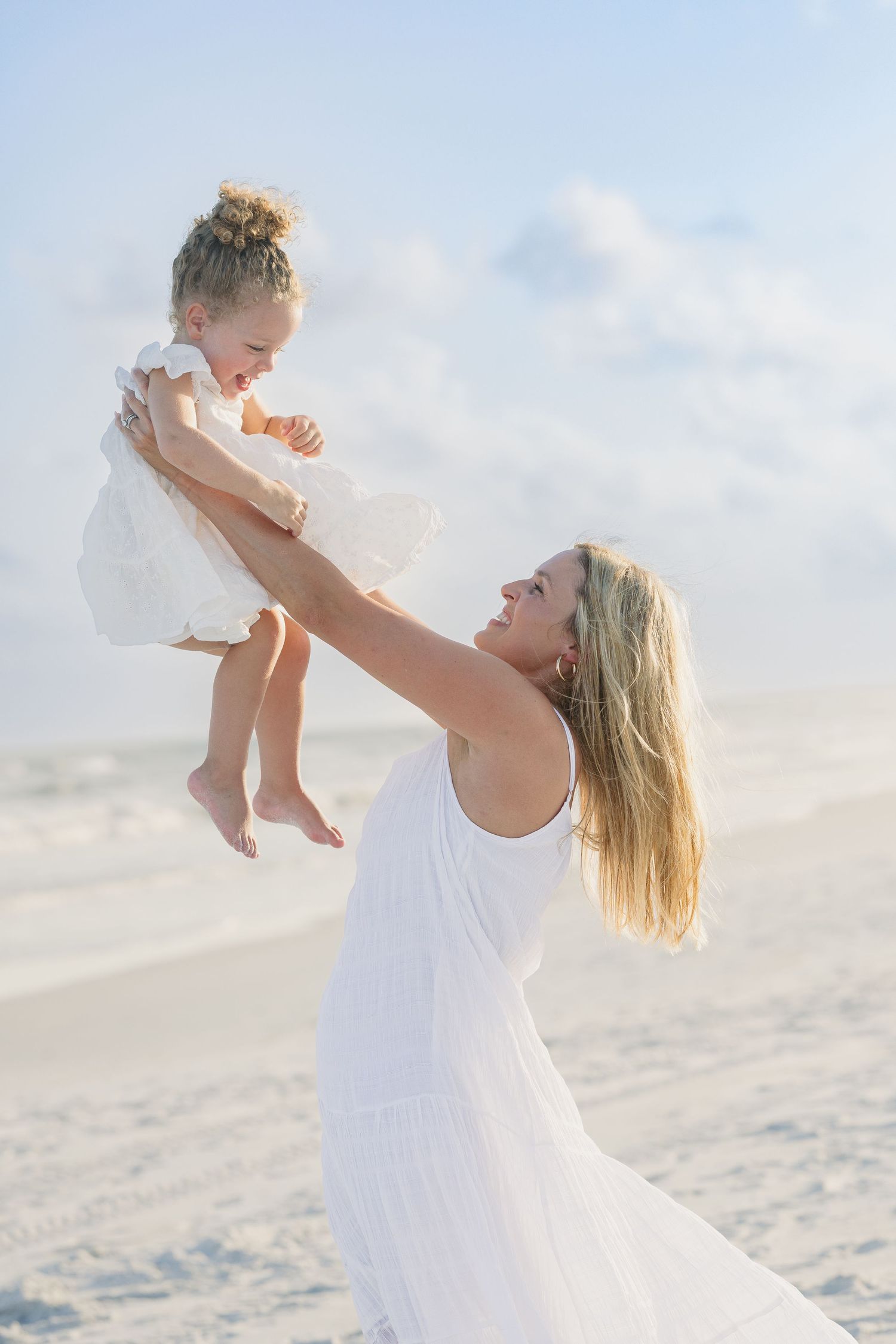 Blonde mother in white dress lifting laughing toddler daughter high in the air on a bright sandy beach.