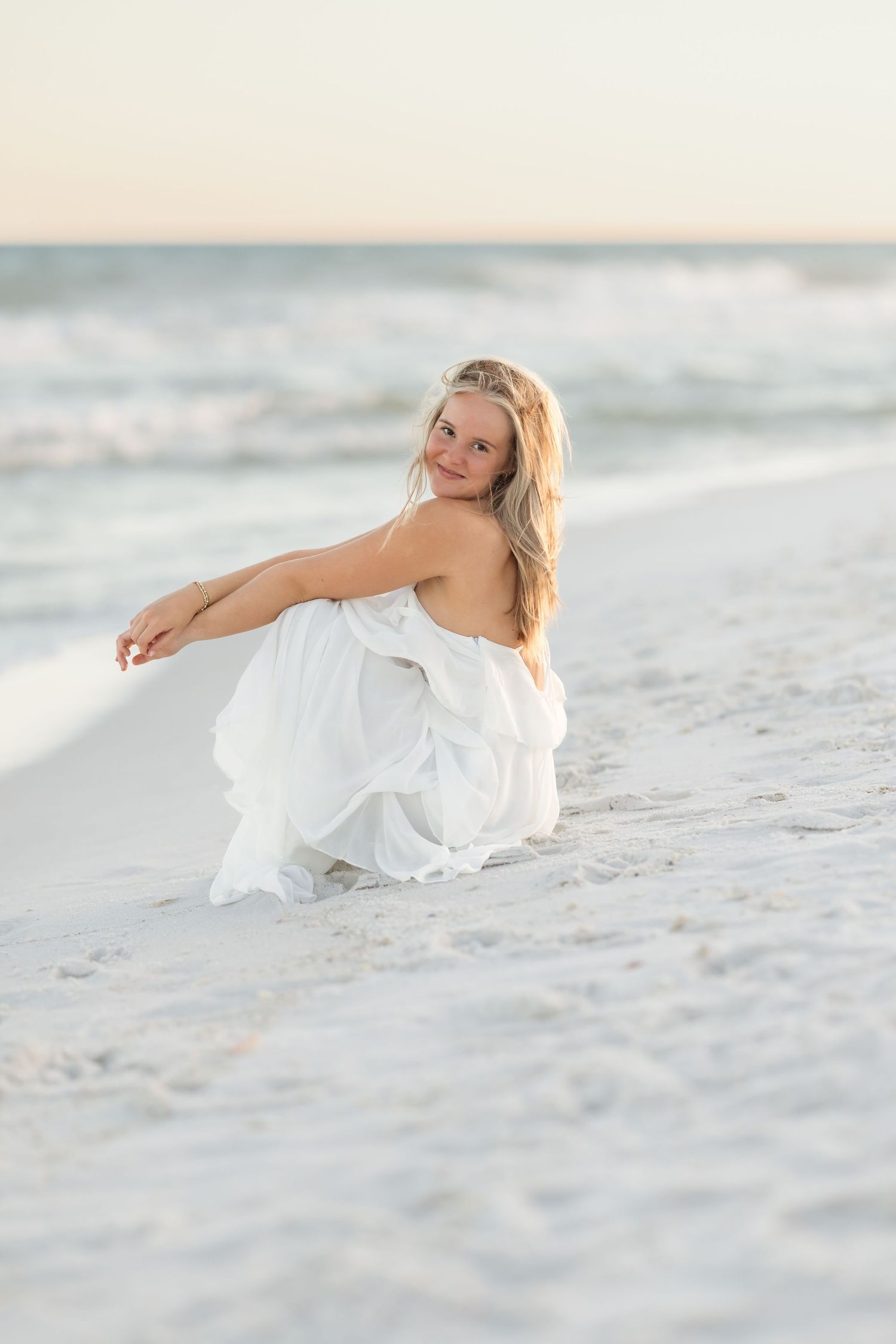 Young blonde woman in white dress sitting on white sand beach with ocean waves in background at sunset.