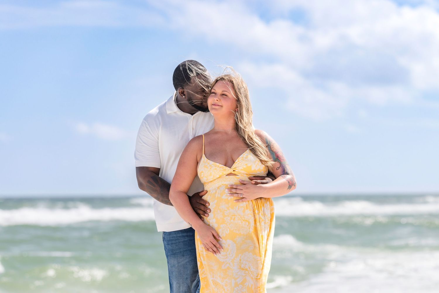 Couple embracing on beach, pregnant woman in yellow floral dress with partner in white shirt, ocean waves behind them.