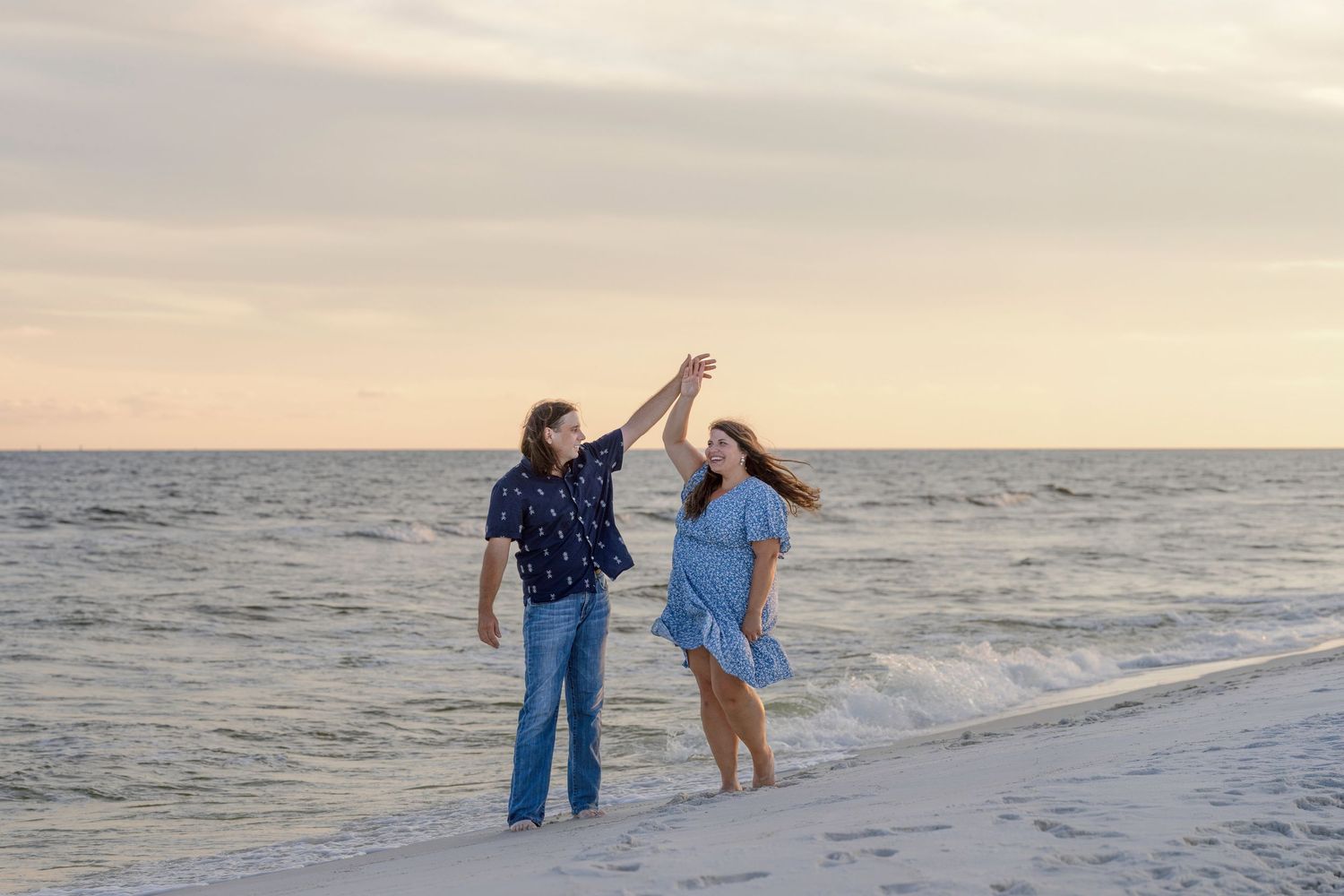 Couple dancing on a sandy beach at sunset, waves in the background, woman in blue dress twirling.