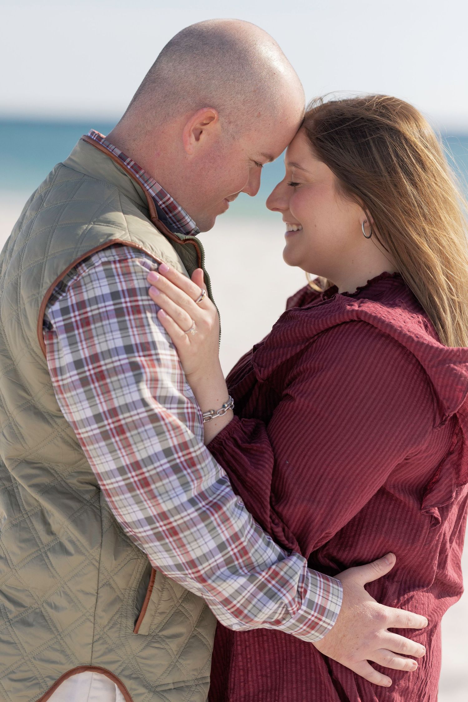 A couple shares a romantic forehead touch on a beach, dressed in plaid and burgundy outfits.